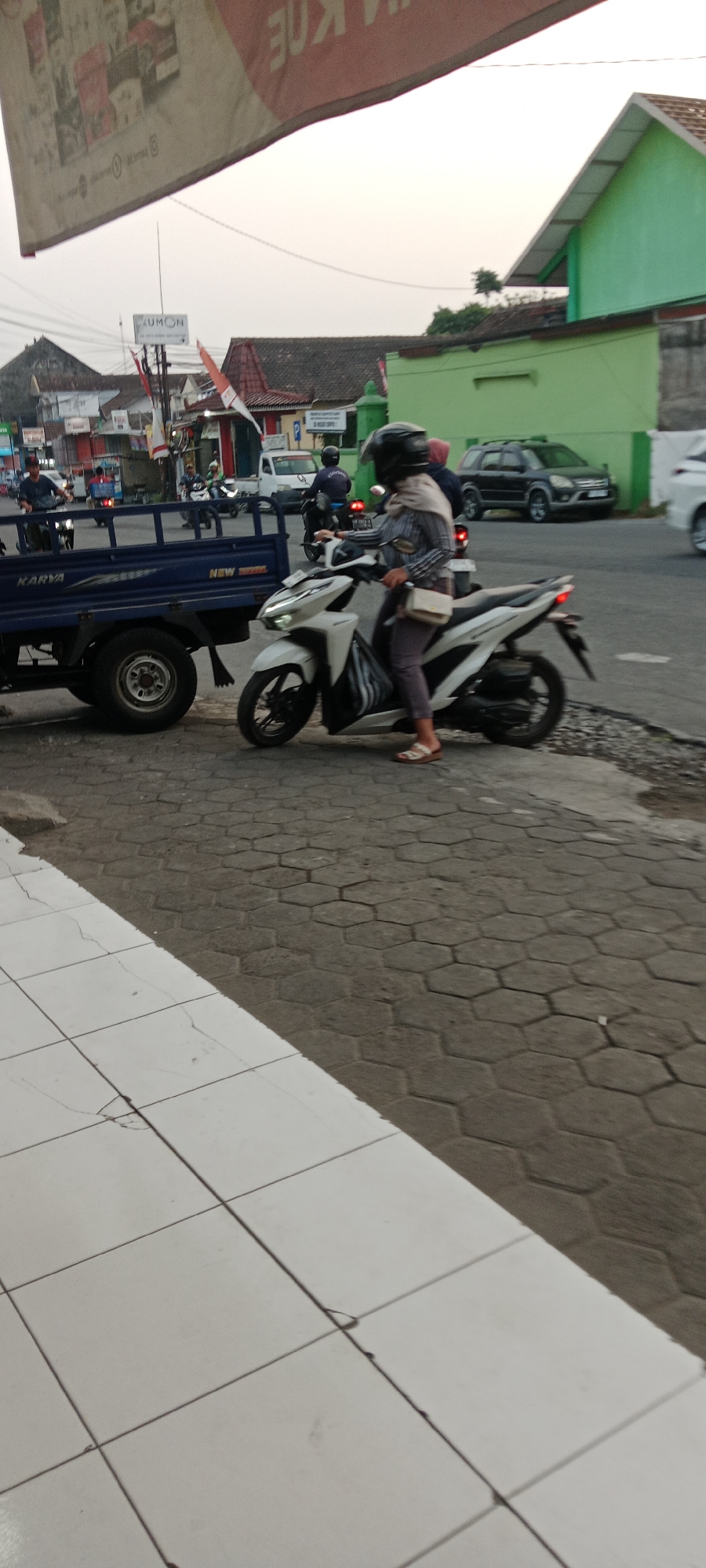 In Maguwoharjo, Indonesia, a person wearing a helmet and a patterned long-sleeved shirt is riding a white motorcycle. The rider is also wearing light-colored pants and sandals and has a light-colored bag slung across their body. The motorcycle has its headlights on. The rider is positioned on the right side of the image. To the left of the motorcycle is the rear section of a blue truck with the word "KARYA" visible on its side. The truck has large wheels. The scene appears to be a street with hexagonal patterned paving stones on the ground, transitioning to a white tiled surface on the left. In the background, there are buildings, including one with a green facade and a white-tiled roof. Other vehicles, including cars and motorcycles, are visible further down the street, suggesting a traffic or urban environment. The sky is overcast.