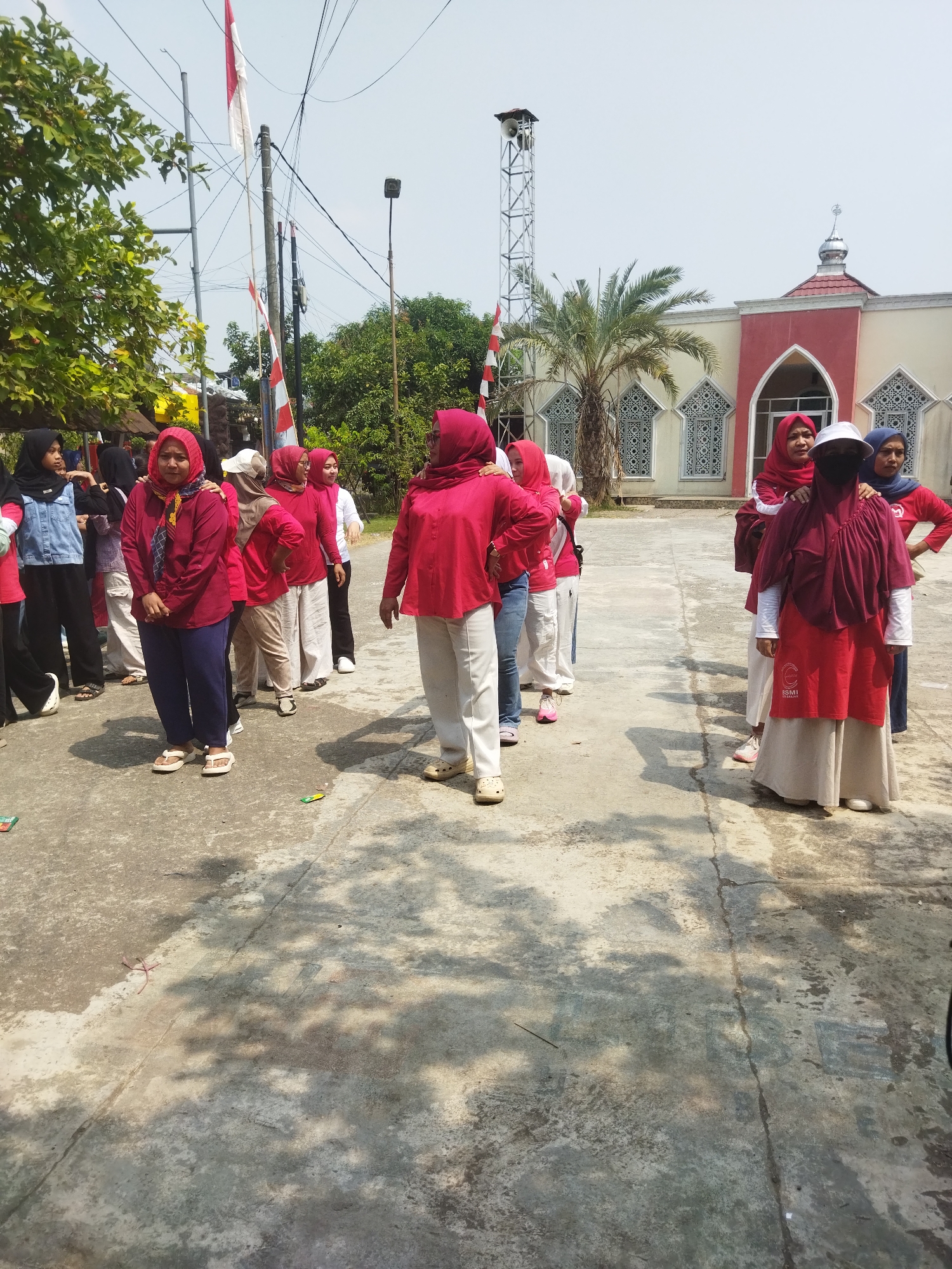 In the city of Banjar, Indonesia, a group of women, predominantly wearing red hijabs and clothing, are gathered outdoors. The scene appears to be in front of a building with architectural elements reminiscent of a mosque, featuring arched windows and a dome. The women are standing in a loose formation on a paved surface, with some engaged in conversation or observing their surroundings. The environment includes trees, flags, and a communication tower in the background, suggesting a public space or community event. The overall atmosphere is casual and social, with the women dressed in modest attire typical of the region.