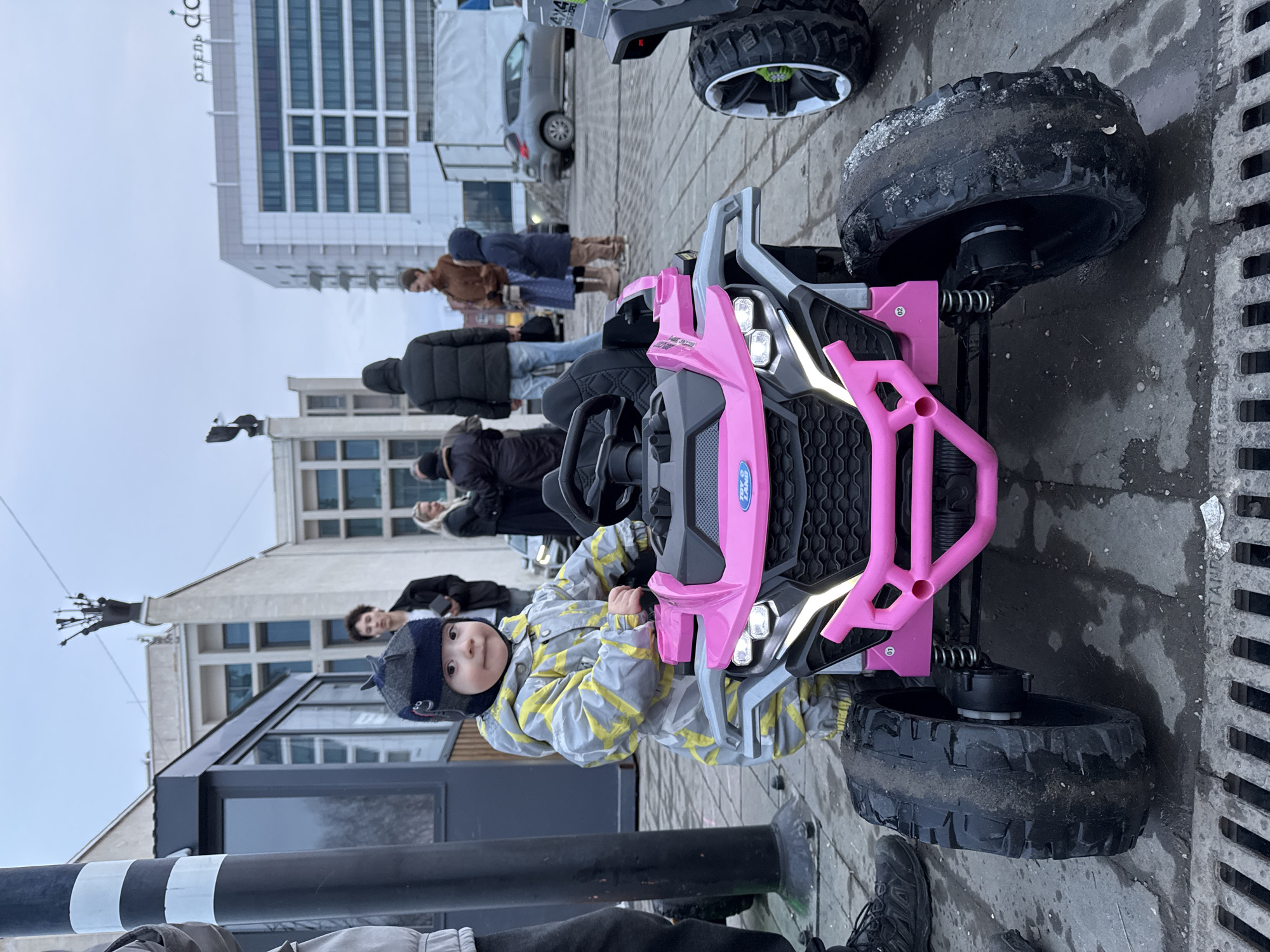 A small child wearing a gray and yellow snowsuit and a dark blue hat sits in a pink toy off-road vehicle on a paved area in Izhevsk, Russia. The vehicle has oversized black tires and distinctive front lights. Several people are standing in the background, including adults wearing winter coats. Behind them are buildings, including a tall, modern structure. A black and white striped pole is visible in the foreground. The paved area appears wet, possibly from melting snow.