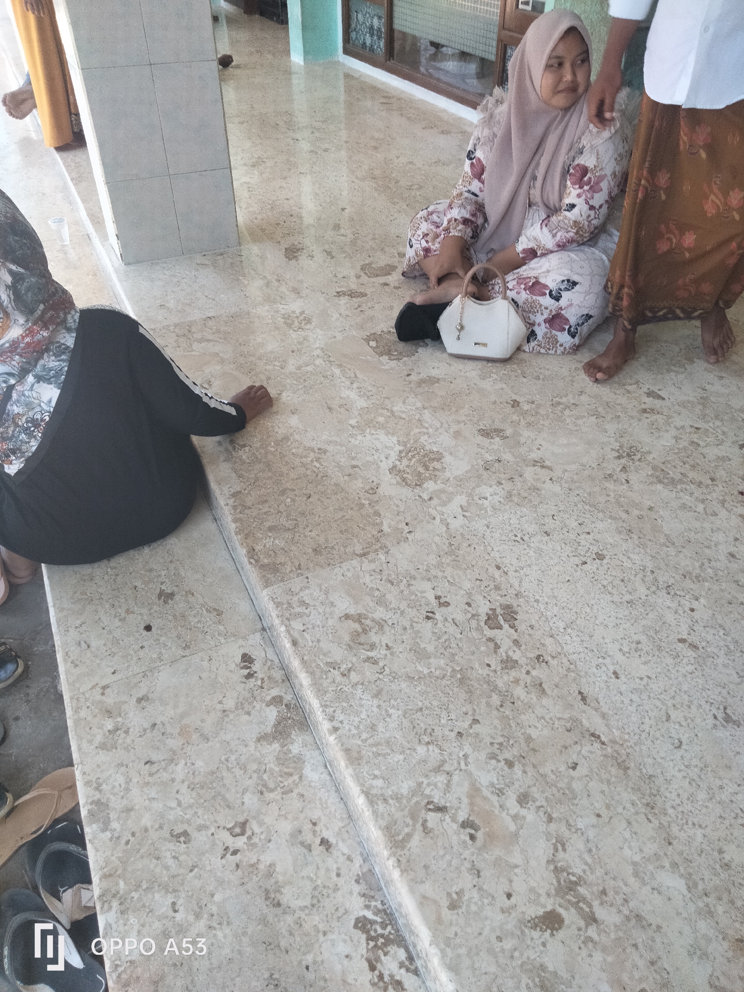 Two people are seated on a light-colored tiled floor. A woman in a hijab and a floral-patterned dress sits with her legs crossed, holding a small white purse. To her right, a person wearing a white shirt and a brown patterned sarong stands, with their right hand resting on the woman's shoulder. Another person is seated on the floor in the foreground, wearing a black garment with white stripes on the sleeves. The environment appears to be indoors, with architectural features and other people partially visible in the background. The floor is made of large, polished tiles with a marble-like pattern.