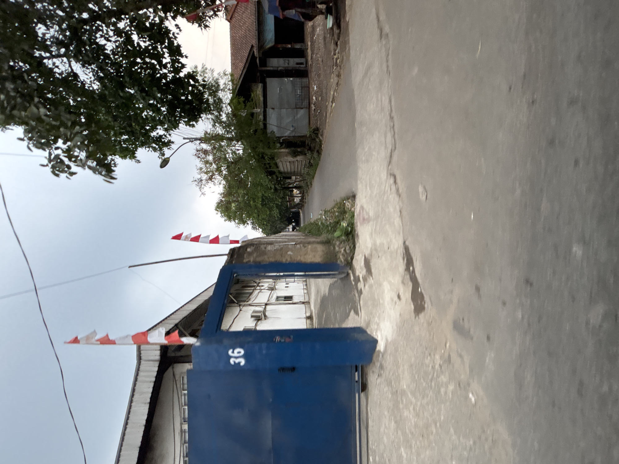 The image shows a street scene in a city, likely in Indonesia given the presence of Indonesian flags. In the foreground, there is a paved road with some debris and water stains. To the left, a large, dark blue metal gate is partially open, revealing a building with a white facade and air conditioning units. The number "36" is visible on the gate. Red and white Indonesian flags are flying from poles attached to the building and to a wall on the right. The street continues into the distance, lined with trees and various buildings, including a structure with a tiled roof and metal shutters. A lamppost is visible among the trees. The sky is overcast. The scene appears to be in a commercial or industrial area.