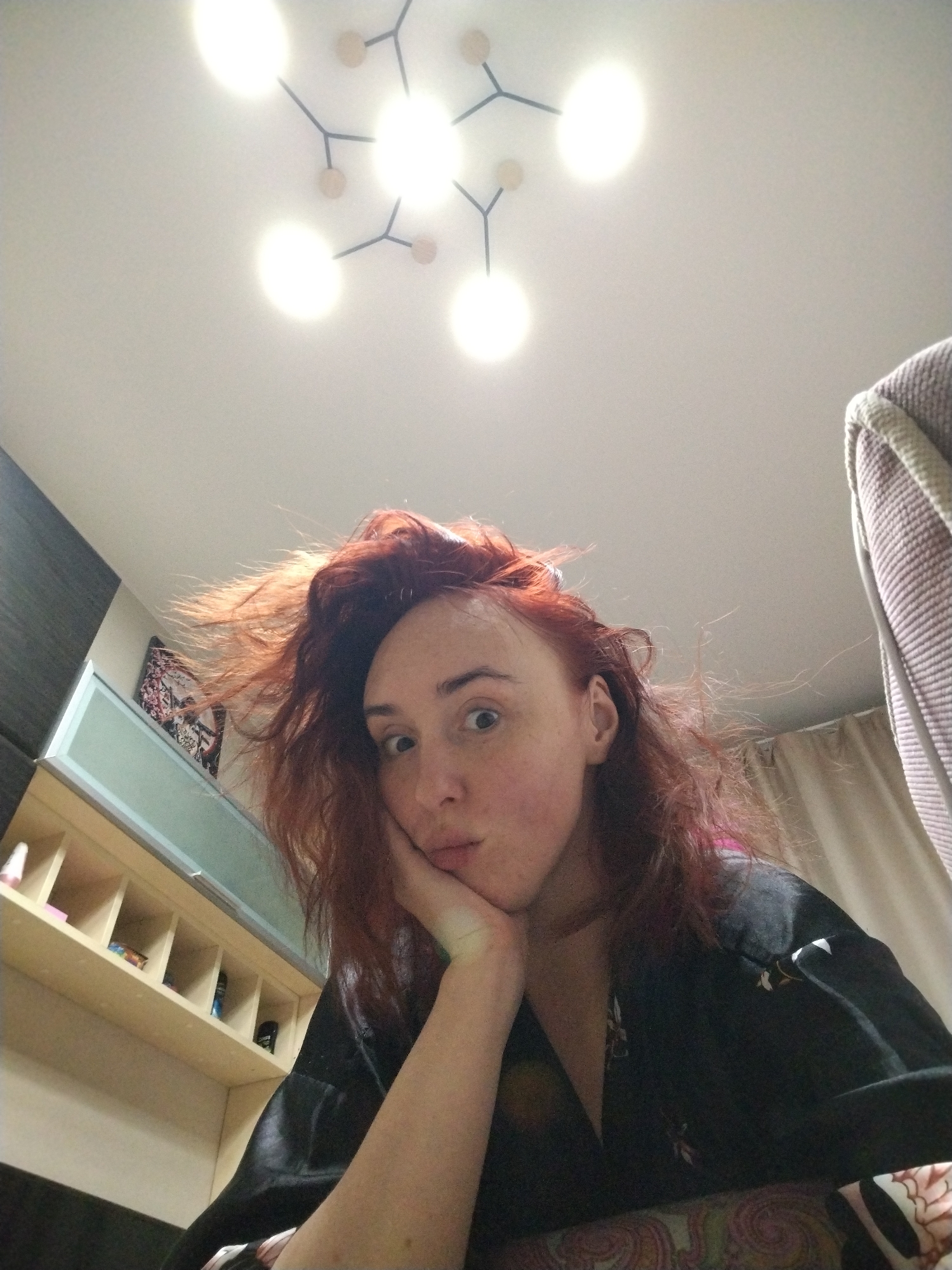 A single individual, appearing female, is depicted in a low-angle selfie captured within an indoor residential room in Izhevsk, Russia. The individual has fair skin, wide eyes gazing directly at the camera, slightly pursed lips, and disheveled reddish-brown hair. Her chin is supported by her right hand. She wears a black garment with subtle light-colored patterns. Above, a white ceiling features a modern black-framed light fixture with at least five illuminated circular bulbs. To the left of the individual, a dark gray wall panel is visible above a light-colored wooden shelving unit containing various small objects. A light blue panel and a partially visible colorful artwork are present higher on the left wall. To the right, a light brown textured textile, possibly a blanket or towel, is draped, with light-colored curtains visible behind it. The individual is positioned against a surface covered by a patterned fabric displaying floral or paisley motifs in shades of brown, pink, and white.