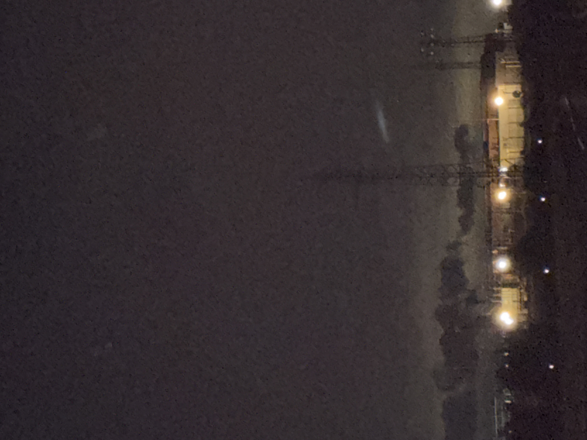 The image depicts an industrial area at night, possibly in Kavarna, Bulgaria, characterized by low visibility and a dark, overcast sky. In the foreground, the landscape appears rugged and uneven, with scattered lights indicating a ground level presence. The middle ground features a complex of industrial buildings illuminated by bright, artificial lights. Tall metal towers, likely communication or industrial structures, are visible, with one prominent tower positioned centrally in the frame. Wisps of smoke or steam rise from behind the buildings, suggesting ongoing industrial activity. A faint, elongated light streak is present in the upper portion of the image, its origin and nature unclear. The overall impression is one of a functional, yet somewhat desolate, industrial landscape under nighttime conditions.