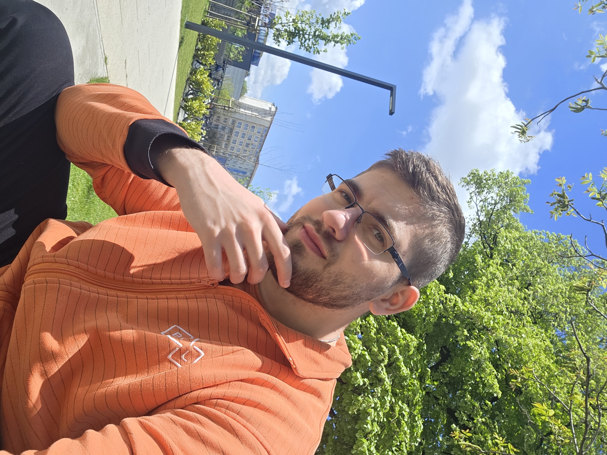 A selfie captured outdoors in Plovdiv, Bulgaria. A man with glasses and a beard, wearing an orange jacket with a white logo, is holding his chin with his right hand. Behind him are green trees and a light blue sky with white clouds. A modern street lamp is also visible, and in the distance, buildings and more greenery are present. The man's posture suggests he's sitting.