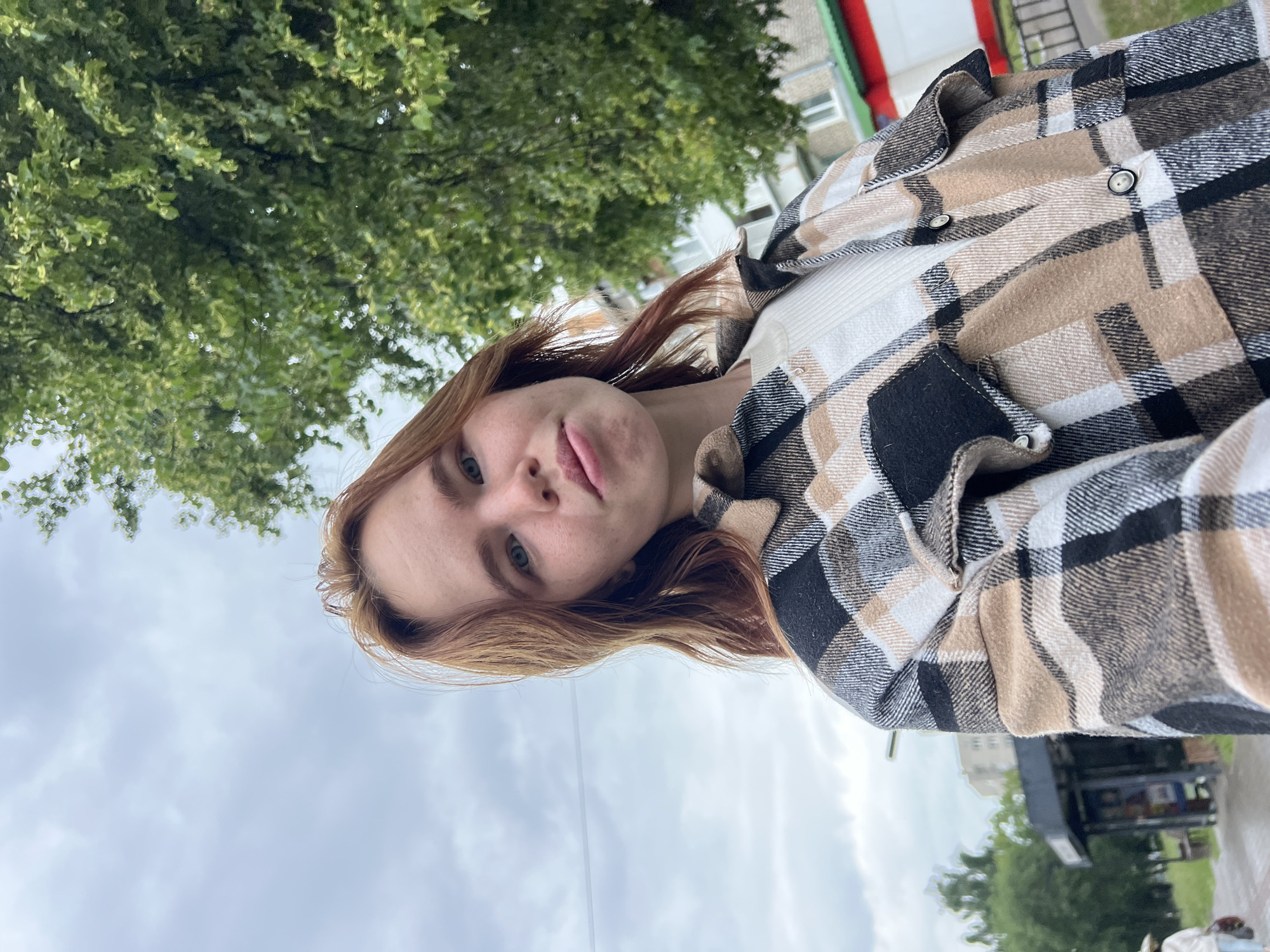 A young woman with reddish-brown hair stands outside, looking towards the camera. She is wearing a plaid shirt over a white ribbed top. The background shows a cloudy sky, green trees, and buildings, suggesting an urban setting. The image appears to be taken in Chaikovsky, Russia, a city known for its industrial presence and proximity to the Kama River.
