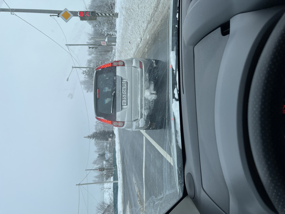 The image captures a snowy winter road scene in Voronino, Russia, from the perspective of a vehicle stopped behind another. In the midground, a silver SUV is stationary on an asphalt road partially covered in snow and slush. The rear of the SUV is soiled with road grime and snow, displaying a Russian license plate "E 353 OA 50". Its red tail lights are illuminated, and a snow-covered tow hitch is visible below the bumper. The vehicle is positioned at an intersection, indicated by a clear stop line on the road. To the right, a utility pole supports a red traffic light and a yellow diamond-shaped 'priority road' sign. Another red traffic light is visible further down the road on the left. The roadside features substantial snow banks, and the background consists of snow-covered bare trees, evergreen conifers, and distant structures under an overcast sky. Utility poles with overhead lines traverse the scene. The foreground shows a partial view of the observer's vehicle dashboard and steering wheel, with snow resting on the interior base of the windshield, which exhibits a visible crack. No individuals are present in the image.