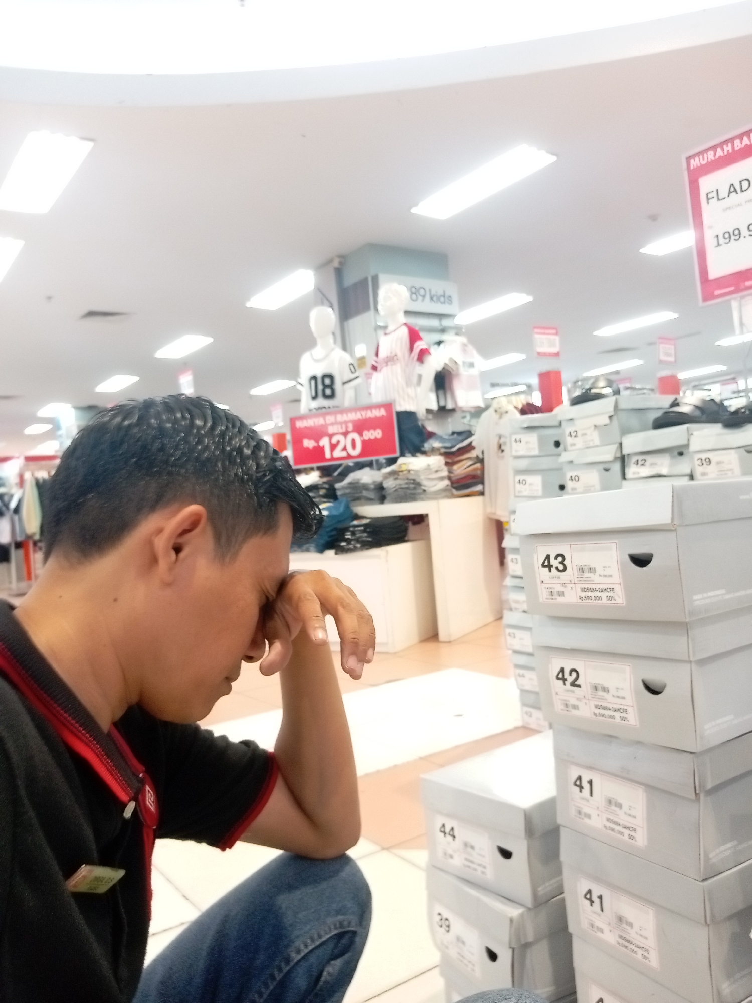 A man is sitting and leaning his head on his hand, with his eyes closed in what appears to be a store. He is wearing a black shirt with red trim and blue jeans. In the background, there are shelves stocked with clothing and shoe boxes. A sign visible in the background reads "FLAD" and "199.9". Another sign indicates a price of "RP 120.000". The environment appears to be a retail store, possibly a department store, with fluorescent lighting. The scene is indoors.