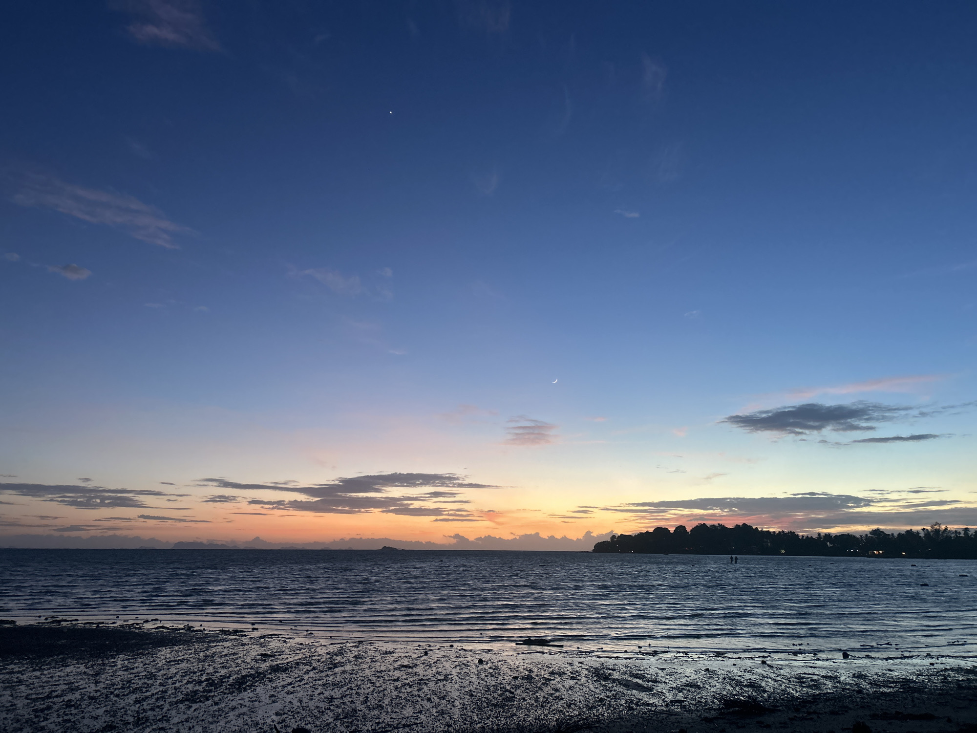 The image depicts a serene twilight scene on the coast of Ko Pha-ngan District, Thailand. A calm body of water, likely the Gulf of Thailand, stretches across the middle of the frame, reflecting the soft hues of the setting sun. The sky transitions from a deep blue at the top to a warm gradient of orange and pink near the horizon, with scattered wispy clouds adding texture.
In the foreground, a wet, reflective shoreline composed of dark sand and small pebbles is visible. The water's surface is rippled, indicating a gentle movement. Across the water, the distant coastline is silhouetted against the colorful sky, with a line of trees and foliage defining the land. Two small figures, appearing to be people, can be seen standing in the shallow water near the distant shore, adding a sense of scale and human presence to the expansive landscape. High in the sky, a faint crescent moon is visible. The overall impression is one of tranquility and natural beauty at the close of day.