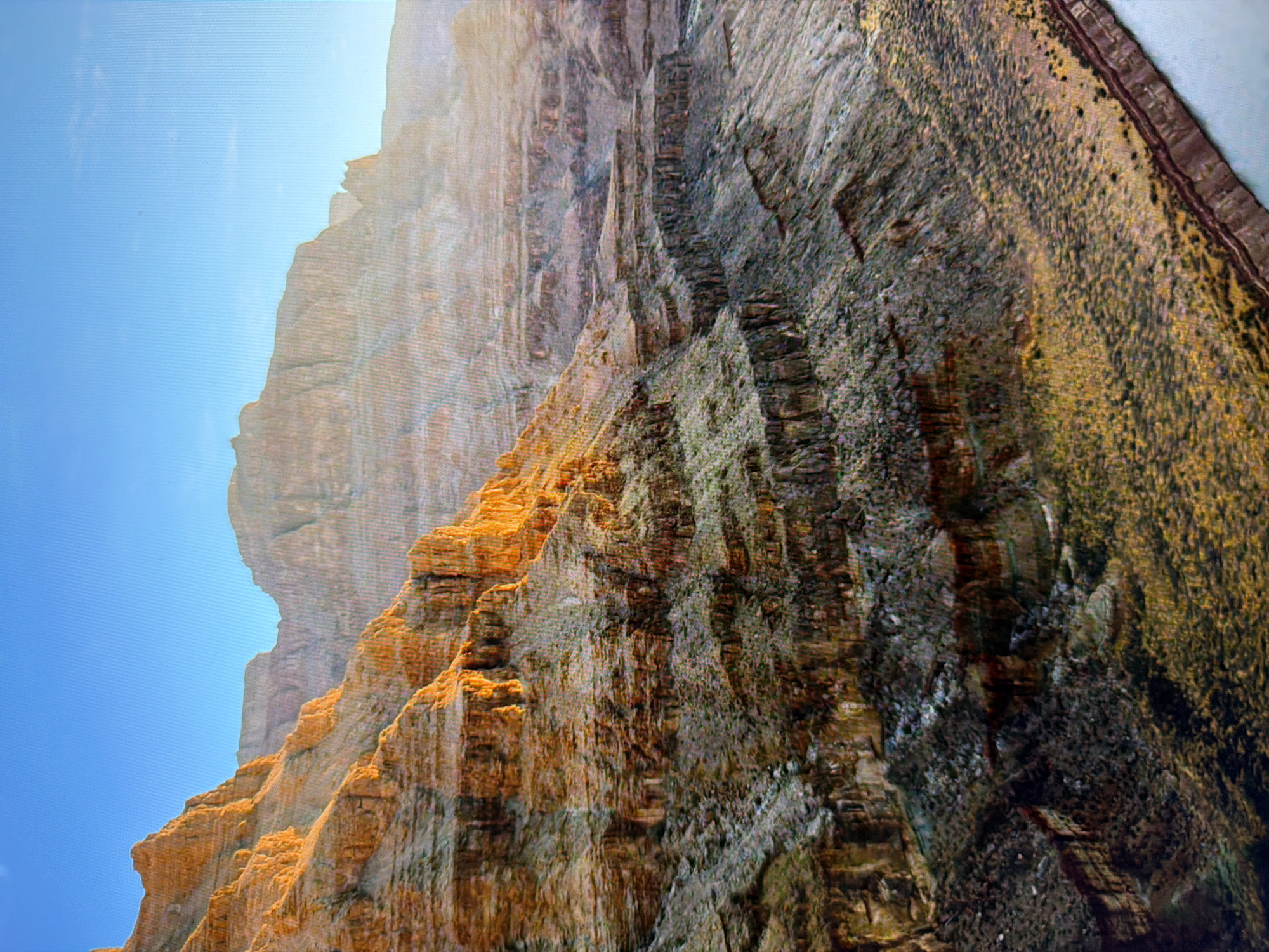 The image depicts a vast canyon landscape bathed in the warm light of what appears to be sunrise or sunset. The sun illuminates the layered rock formations on the left side of the frame with a golden hue, highlighting the textures and shadows. The canyon walls exhibit distinct horizontal stratification, showcasing geological history. Sparse vegetation, a mix of scrub and grasses, covers the slopes and the canyon floor. In the lower right corner, a body of water, likely a river, is visible, its surface reflecting the light. The sky is clear and a bright blue. The scene is a natural landscape, characteristic of a grand geological formation like the Grand Canyon. There is no indication or any visual element that would place this scene in Kyiv, Ukraine, as the geography and geological features are not consistent with that location.