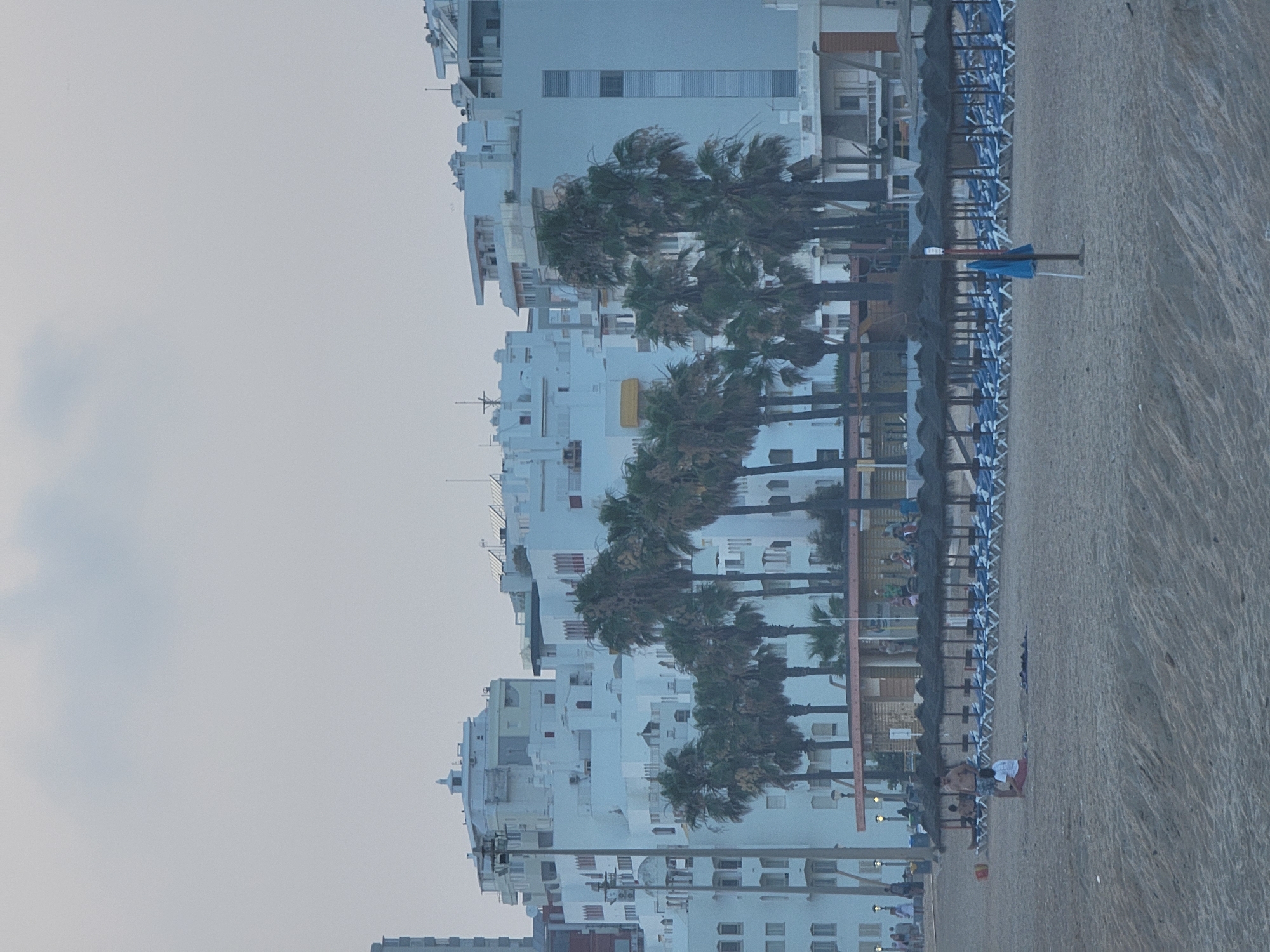 The image depicts a beach in Quarteira, Portugal. The scene is dominated by a row of white buildings with architectural elements such as balconies and decorative trim. Palm trees are interspersed in front of the buildings. In the foreground, the sandy beach is lined with numerous empty lounge chairs and straw umbrellas, suggesting a resort area. A few people are visible on the beach and near the buildings. One man is standing on the beach, and another person is sitting on the sand. A blue umbrella is planted in the sand, possibly for shade. The sky is overcast, and the overall lighting suggests it is either early morning or late evening.