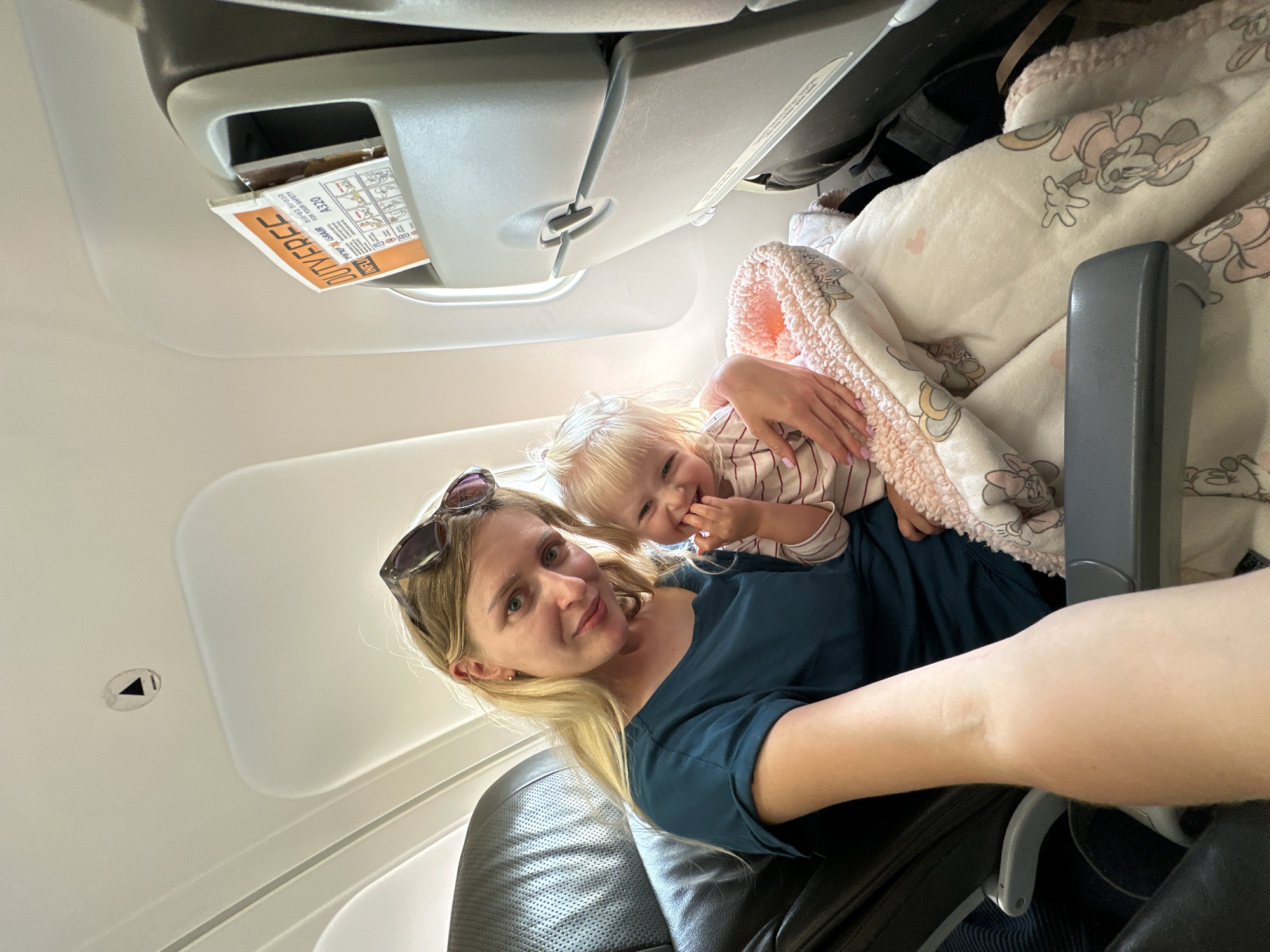 A blonde woman and a baby are seated on an airplane. The woman is wearing a dark teal shirt and sunglasses are perched on her head. She is holding the baby, who is wearing a striped shirt and is wrapped in a pink and white blanket with Mickey Mouse patterns. The baby is smiling and has its fingers near its mouth. The airplane cabin is visible, with seats and a safety card in a pocket on the seat in front. The image appears to be taken in Yehud Monosson, Israel.