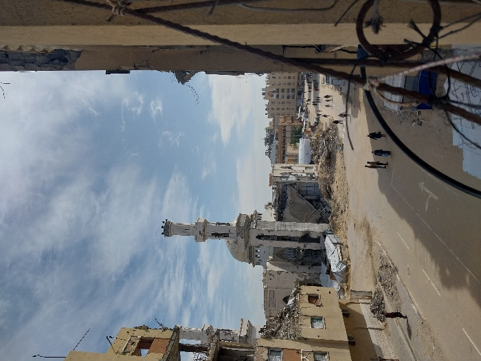 The image depicts a scene of extensive urban destruction in Khan Yunis, Palestinian Territory, viewed from an elevated position. The foreground right shows a portion of a damaged building wall with exposed rebar and wiring. To the left, a multi-story building is severely damaged, with its upper floors and interior exposed and rubble scattered around its base.
In the mid-ground, a prominent white mosque with a tall, slender minaret stands amidst widespread devastation. The mosque structure itself is heavily damaged, with significant portions of its walls and roof missing. The minaret shows structural damage near its base and top. To the right of the mosque, large piles of rubble and debris, including twisted metal and corrugated sheets, mark the site of collapsed buildings.
A paved street runs through the center of the scene, with a white arrow road marking. Numerous pedestrians are visible on the street. A group of approximately five individuals is in the mid-ground, walking away from the camera. Further down the street, an additional ten to fifteen people are dispersed, also moving along the road. One solitary figure walks on the left sidewalk towards the camera.
In the background, beyond the immediate destruction, more multi-story residential buildings are visible, some appearing less damaged, along with a large white tent-like structure. The sky is partly cloudy with scattered wispy clouds. The overall environment is characterized by widespread structural damage, rubble, and ongoing pedestrian movement.