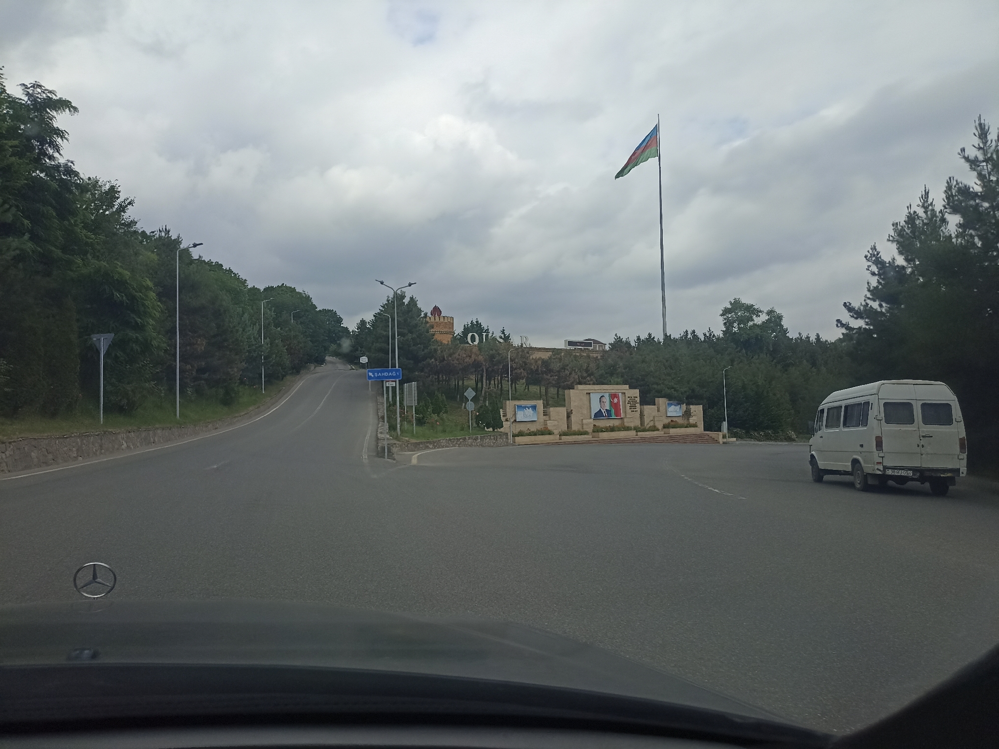 The image shows a road with a white van driving on the right side. In the background, there is a monument with portraits of two men and a flag of Azerbaijan. The monument is located in the city of Qusar, Azerbaijan, which is known for its mountainous terrain and beautiful natural scenery. The sky is cloudy, and the trees are lush green, suggesting it is summertime. A Mercedes-Benz logo is visible on the hood of the car from which the photo was taken.