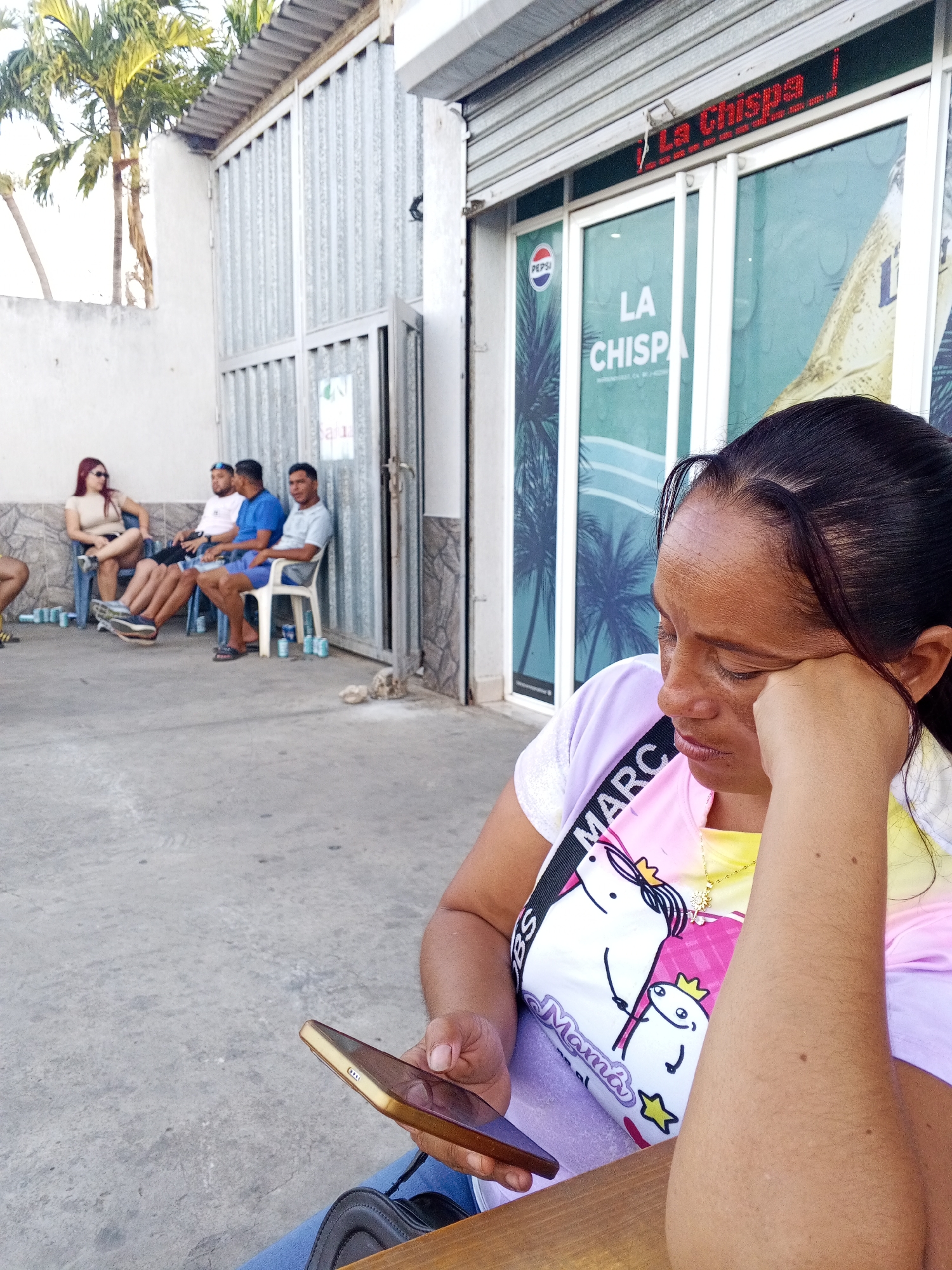 A woman is in the foreground holding a cell phone, her head resting on her hand. Behind her, three men and a woman are sitting on chairs in front of a building, likely a business, which has a sign that reads "La Chispa." The city location is Unknown, Unknown. The environment includes buildings, palm trees, and a concrete surface.