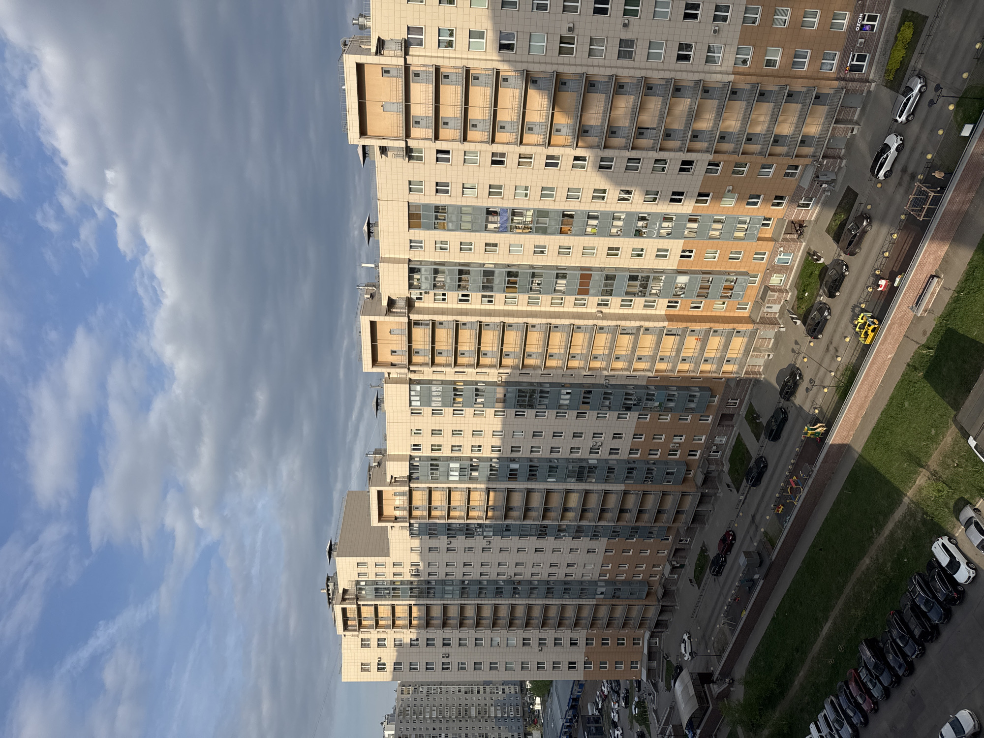 The image shows a high-angle view of a residential area in Saint Petersburg, Russia. Multiple tall apartment buildings with beige and gray facades are visible, casting shadows onto the street below. The street is lined with parked cars, a narrow strip of grass, and a pedestrian walkway. There are also some vehicles moving along the road. A blue sky with clouds is partially visible in the background.