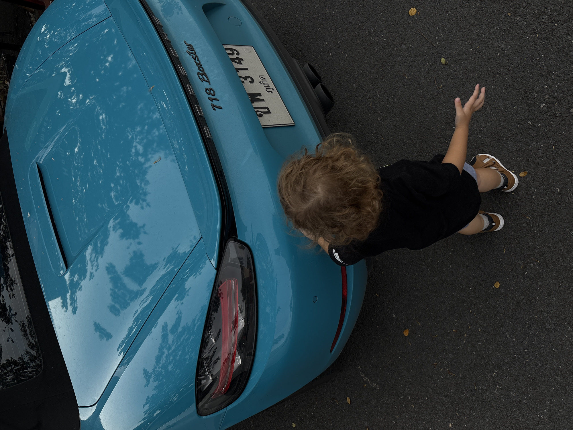 A young child with curly blonde hair, wearing a black t-shirt and light-colored shorts, stands near the rear of a bright blue Porsche 718 Boxster. The child's right arm is extended upwards, with their hand open. The car's rear is visible, featuring a license plate with Thai characters and the number "3149". The car has dual exhaust pipes.
The scene appears to be outdoors, on an asphalt surface, possibly a street or parking lot. A few fallen leaves are scattered on the ground, suggesting an outdoor setting. The lighting indicates it is daytime, with soft light and reflections on the car's polished surface. The overall mood seems casual and playful, with the child interacting with the car.
Visible text on the car includes "718 Boxster" and a Thai license plate. The license plate has characters that translate to "Chang Wat Phuket," indicating the vehicle is registered in Phuket, Thailand, although the image context places it in Choeng Thale.