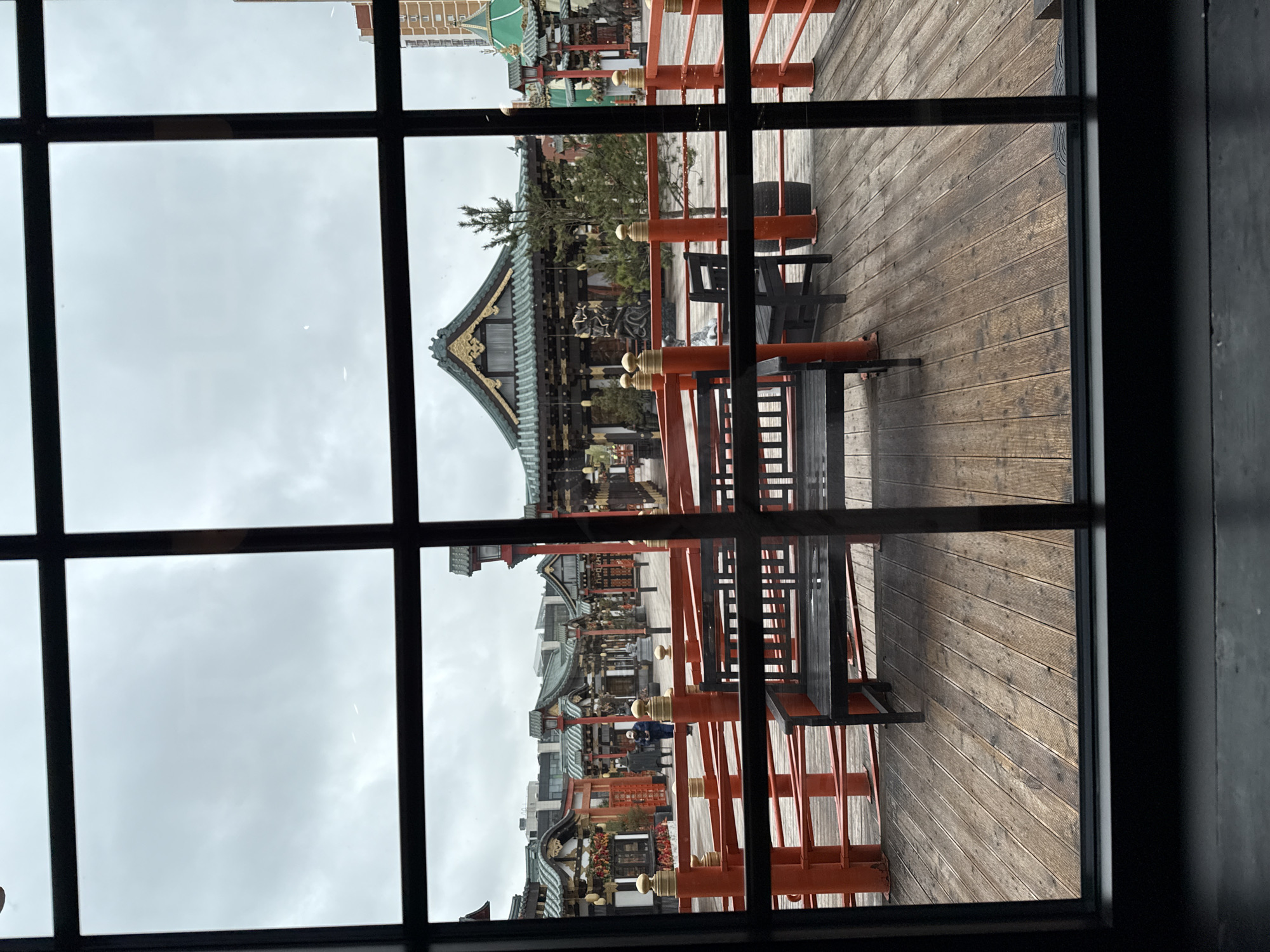 The image captures an outdoor scene with distinctive East Asian architecture, viewed through a window featuring a dark, prominent grid pattern. The perspective is from an interior space looking out onto an exterior area.
The sky is overcast and grey, indicating a cloudy or dim day. Immediately outside the window, a weathered wooden deck or platform extends across the lower and right portions of the frame. Bright orange-red railings, adorned with golden finials, are a prominent feature on this deck, suggesting a balcony or a protective barrier. Several dark, simple tables or benches are interspersed among these railings.
Beyond the deck, a series of traditional East Asian buildings dominate the mid-ground. These structures showcase intricate, curved roofs with dark green tiles and ornate golden decorations. One prominent building has a particularly elaborate roof design and is partially framed by sparse greenery, possibly a small tree or shrub. Further in the distance, a modern building with a lighter facade can be seen peeking out above the traditional architecture.
A lone individual, dressed in a dark blue or black outfit, is visible standing near the base of one of the traditional buildings in the mid-ground. Their presence suggests the area is either a public space, a cultural site, or a residential complex. No specific activity is discernible from the person's posture. The scene evokes the ambiance of a cultural park, temple grounds, or a historically themed district. No text is visible in the image.
