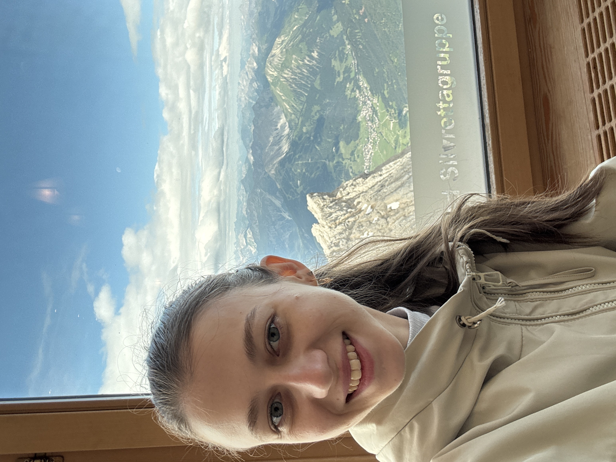 A young woman with brown hair and blue eyes smiles at the camera, her face filling the left side of the frame. She is wearing a light-colored jacket over a white turtleneck. Behind her, a large window offers a panoramic view of the Alps, with lush green valleys dotted with small villages and rugged, rocky peaks reaching towards a bright blue sky with scattered clouds. The text "Silvrettagruppe" is visible on the window, indicating the mountain range. The scene suggests the woman is at a high vantage point, likely a tourist attraction or a mountain hut in the Austrian Alps, possibly near Ehrwald.