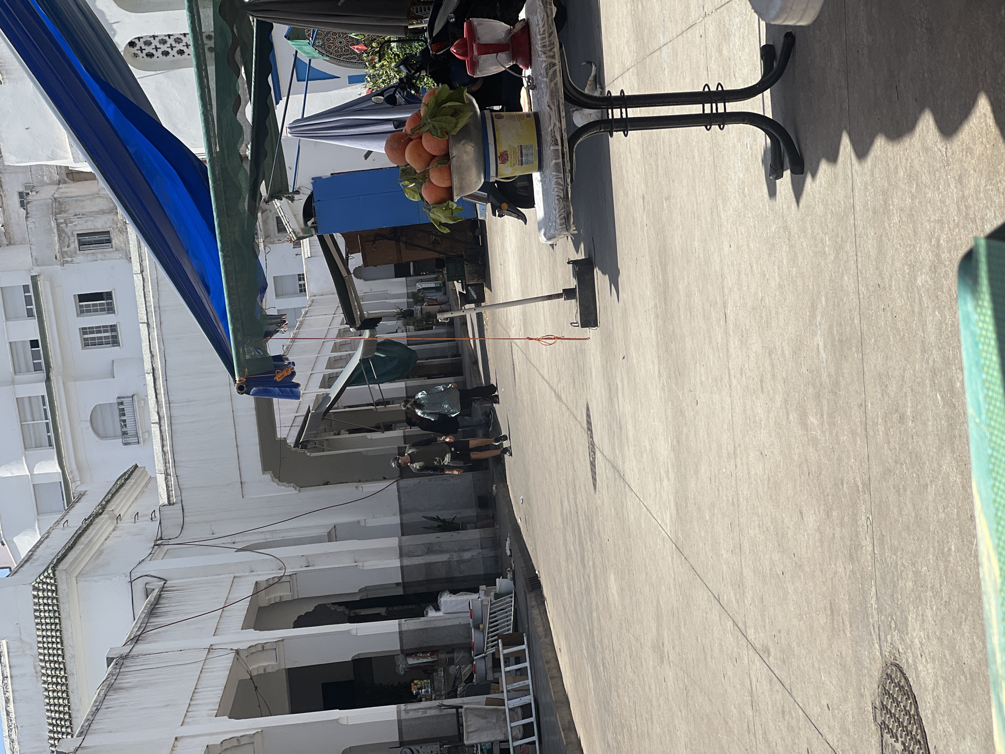 The image captures a vibrant street scene, likely in a market area in Casablanca, Morocco, under bright daylight. The main focal point in the midground is an outdoor vendor's setup, featuring a black metal table whose surface is covered with a white sheet or plastic. On the table, a silver bowl overflowing with fresh oranges, many still bearing green leaves, is prominently displayed. Behind the oranges, a red and white citrus juicer is visible, suggesting a fresh juice stall. A blue kiosk or stall structure stands behind this setup.
The ground is a light-colored concrete or pavement, showing signs of wear and tear, with strong shadows cast by the table and other unseen elements, indicating bright sunshine. A circular manhole cover is visible on the right side of the pavement. In the background, several white multi-story buildings dominate the landscape, characterized by traditional Moroccan architecture with small windows and some decorative elements. Power lines or cables are strung along these buildings. A blue tarp or awning extends across the upper left of the frame, providing shade, and another dark green awning is also present.
A few individuals are scattered in the background, engaged in daily activities, walking or standing near the stalls. One person wears a dark jacket, and another a lighter top. Further back, a ladder is seen leaning against a building. The overall atmosphere suggests a typical bustling street or market environment. The time of day is clearly sunny, likely morning or afternoon, given the distinct shadows. No legible text is discernible within the image.