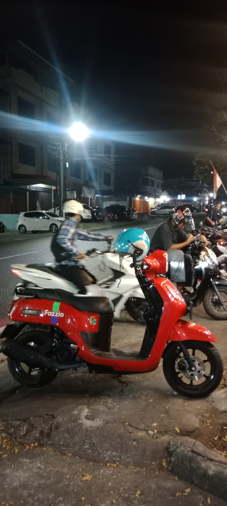 The image shows a street scene at night in Tanjung Pinang, Indonesia. In the foreground, a red Yamaha Fazzio scooter is parked on the side of the road. The scooter has the Fazzio logo on its side, along with a green and blue stripe. A blue helmet with white markings rests on the seat. Behind the red scooter, a white scooter is visible, with a person wearing a checkered long-sleeved shirt and a white helmet riding it. Further back, several cars, including a white hatchback, are parked along the street. Buildings with illuminated windows line the street on the left side. The road surface is asphalt, with some cracks and fallen leaves. A lamppost with a bright light is present on the left side of the image, casting light and lens flares. The overall atmosphere is that of a typical urban street at night, with artificial lighting illuminating the scene.