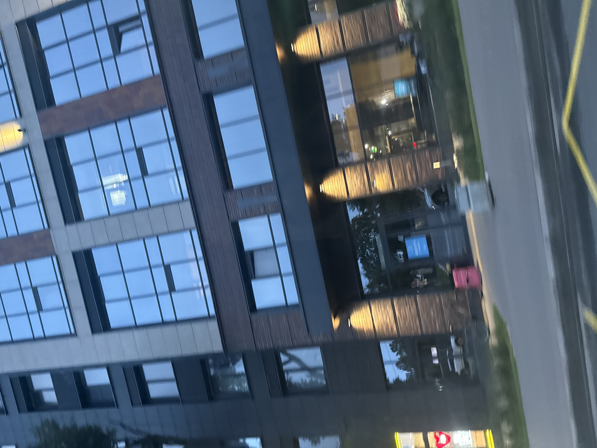 The image captures a street-level view of a modern, multi-story building with a dark brick facade and large, reflective windows. The building appears to be commercial or residential, with several levels visible. The windows reflect the sky and surrounding trees, suggesting an outdoor setting.
At street level, there is an entrance area with warm lighting illuminating the facade. A white SUV is parked near the entrance. To the left, a brightly lit sign with a red heart shape is visible, indicating a commercial establishment. A pink trash bin is placed near the entrance on the right. The sidewalk area shows some greenery, and a concrete barrier is visible in the foreground.
The scene is somewhat tilted, suggesting the photo was taken from a moving vehicle or at an angle. The overall lighting suggests it is either dusk or an overcast day.
Given the architectural style and the prevalence of similar modern buildings with large windows and brick facades, this image could be located in a contemporary urban development area of Saint Petersburg, Russia. Many new residential complexes and business centers in districts like Primorsky, Kalinino, or Nevsky, particularly those developed in the last 10-15 years, feature this type of design. The presence of a car and pedestrian access points points towards a mixed-use or residential area with commercial storefronts.