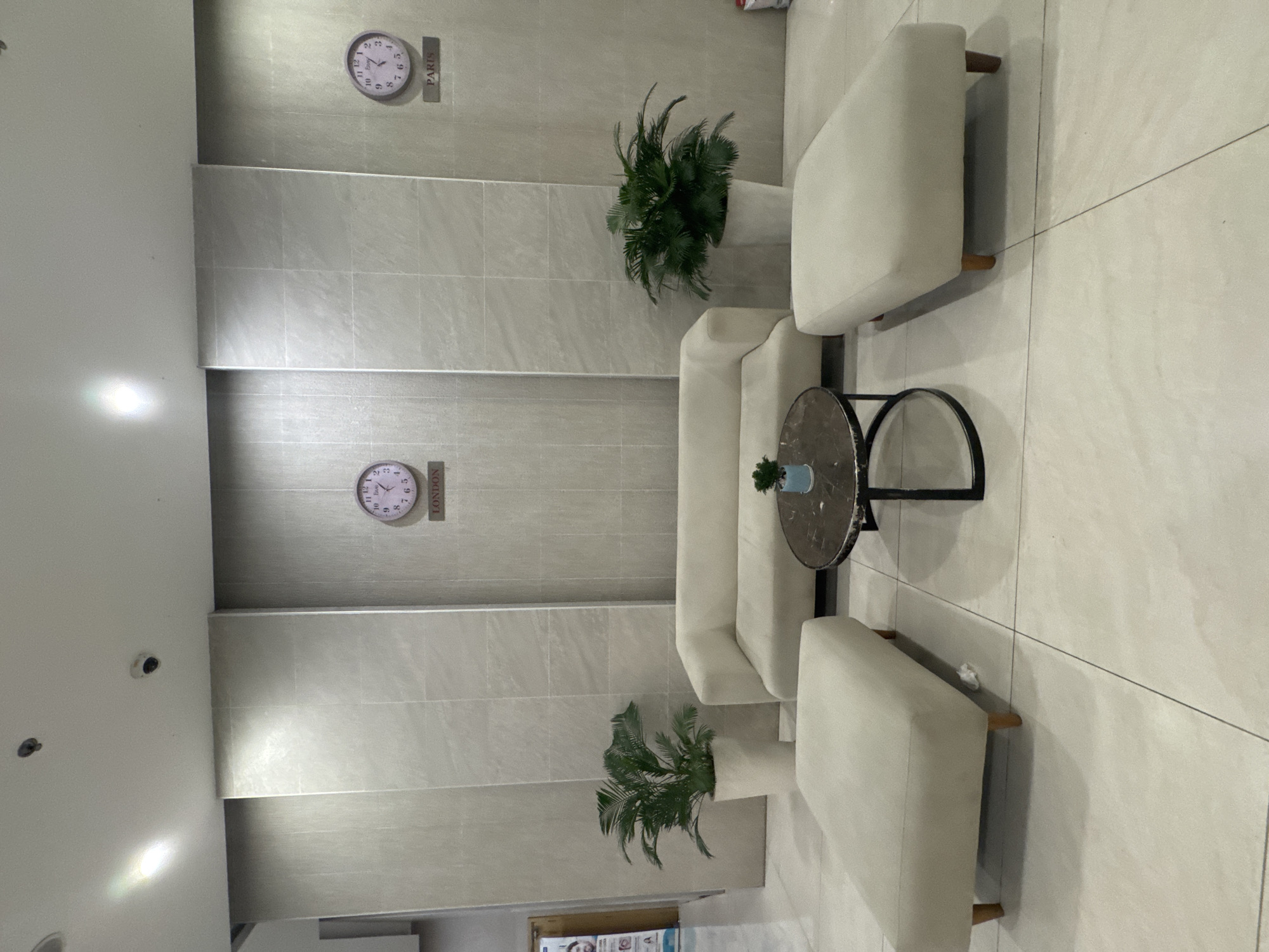 The image shows a waiting area with modern, minimalist furniture. In the center, there is a long, off-white couch. In front of the couch is a round coffee table with a dark, patterned top and a black metal base. Two identical off-white ottomans, one on either side of the coffee table, flank the couch. In the background, the wall is covered in light-colored, large-format tiles. Two clocks are mounted on the wall; one labeled "LONDON" and the other "PARIS." There are also two potted plants, one on the left and one on the right, with lush green foliage in white pots. The floor is tiled with large, light-colored tiles. The scene is well-lit, possibly by overhead lighting, and appears to be an indoor waiting area. There are no people visible in the image. The location is described as being in Bac Nha Trang Ward, Vietnam.
