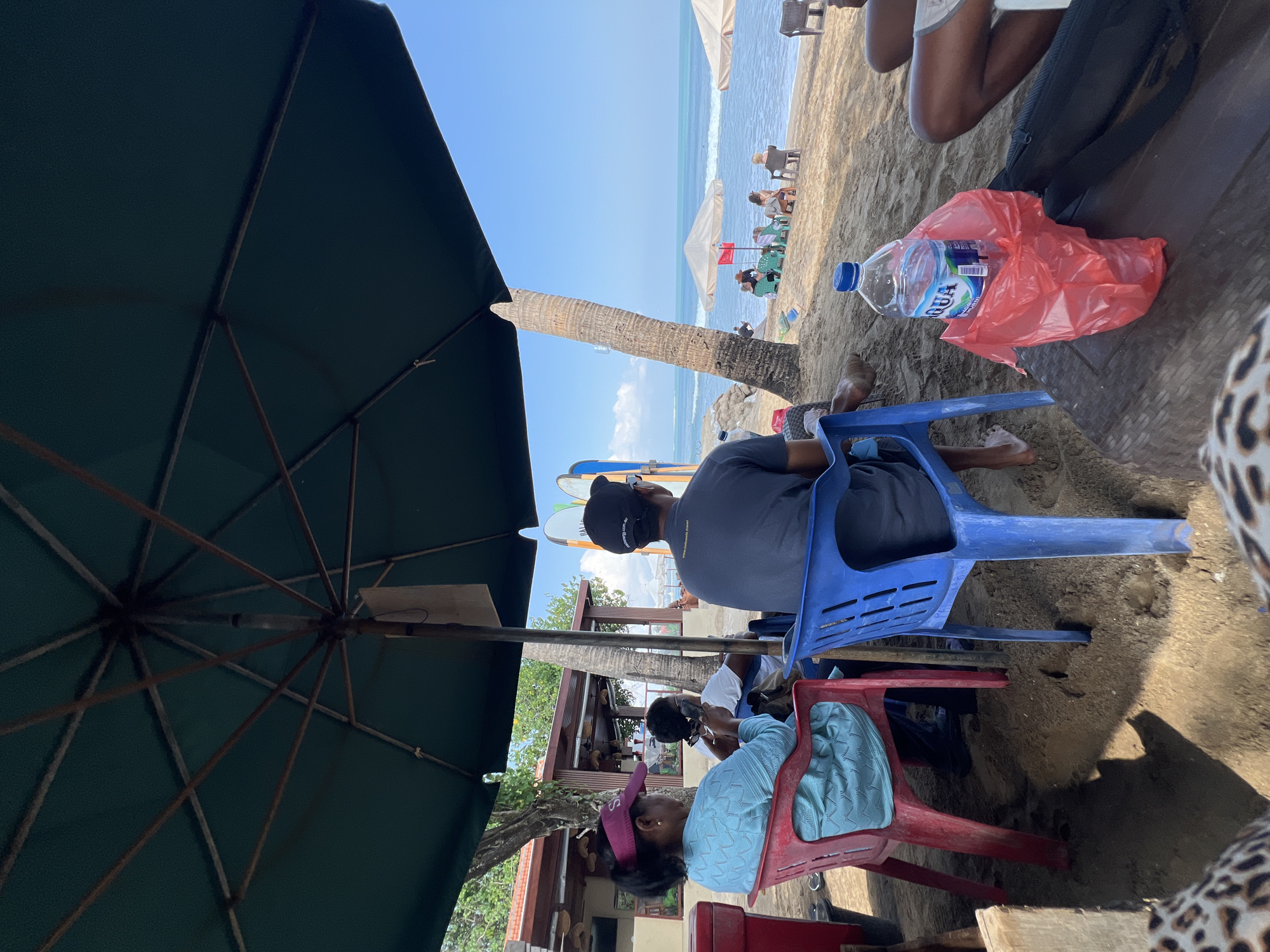 The image captures a scene at Kuta Beach, Indonesia, under a large dark green umbrella. In the foreground, three individuals are seated on plastic chairs, facing away from the camera and towards the ocean. On the left, a woman wearing a pink visor and a light blue patterned top is seated on a red chair, looking down at her phone. Next to her, on a white chair, a person in a white shirt is also looking down. To the right, a man in a dark t-shirt and a black baseball cap is seated on a blue chair, his gaze directed towards the sea.
Further back, several other people are visible on the sandy beach, some seated under umbrellas and others walking. Two surfboards are propped up vertically behind the people in the foreground. The ocean stretches out to the horizon under a clear blue sky, with gentle waves rolling onto the shore. A palm tree stands to the right of the foreground group, and a building with a brown roof is visible in the background to the left, suggesting a beachfront establishment. A plastic bottle of Aqua water and a red plastic bag are placed on a brown surface in the immediate foreground. The overall atmosphere is relaxed, suggesting a leisurely day at the beach.