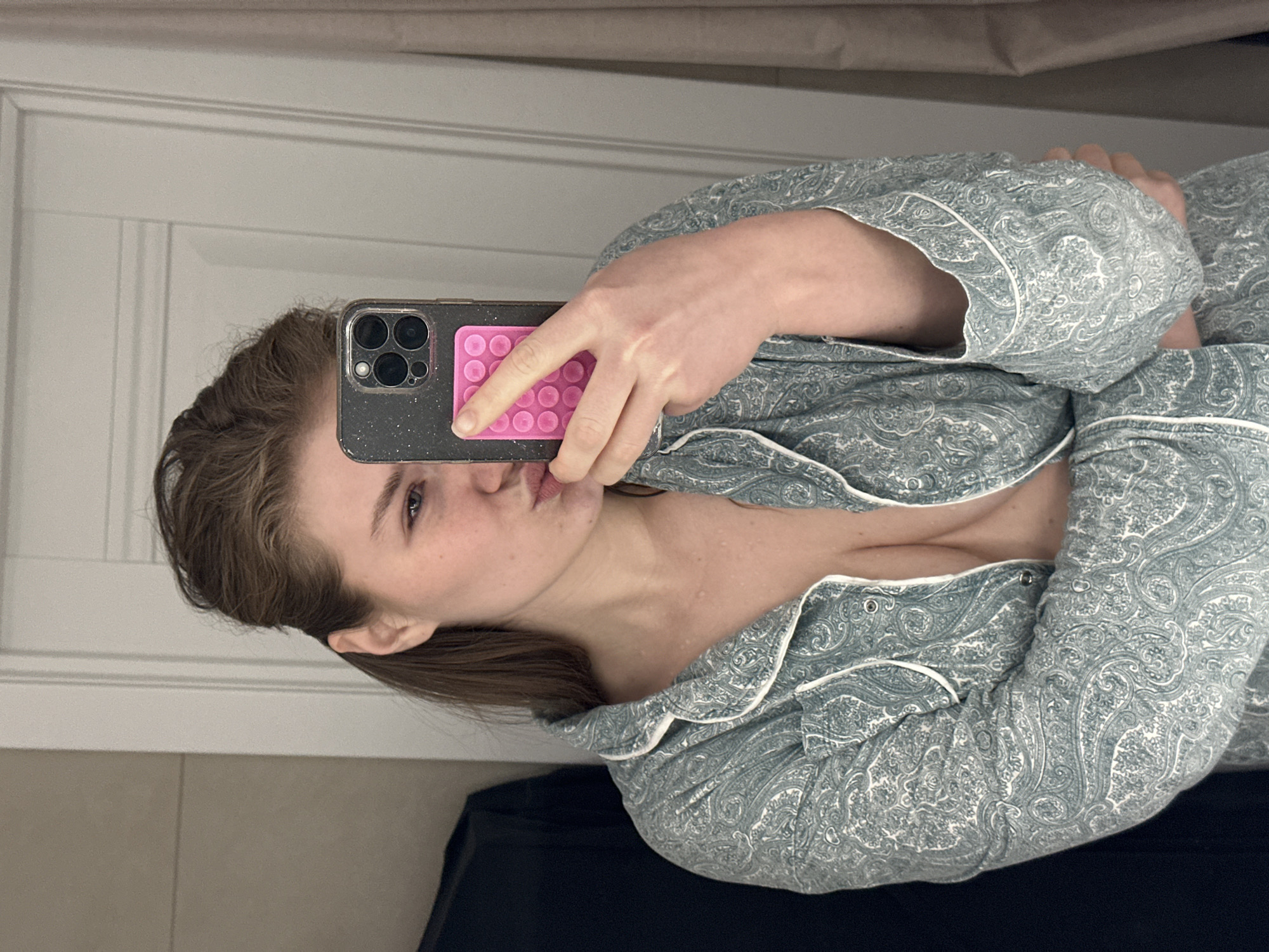 A young adult female is depicted in a close-up mirror selfie, framed from the chest up against an indoor background. She has light skin, brown hair, and one visible blue eye, with her lips pursed in a 'kissing' expression. She wears a collared pajama top featuring a green-blue paisley pattern with white piping; the top is unbuttoned, revealing her cleavage. Her left hand holds a dark-colored smartphone, possibly an iPhone model with multiple rear cameras, which is encased in a glittery clear cover. A bright pink accessory, resembling a 'pop-it' fidget toy with circular indentations, is attached to the back of the phone. The background consists of a white paneled door, a light-colored wall, a dark garment or item to the left, and a beige curtain or drapery to the right. The image does not provide visual cues to confirm a specific location in Kazan, Russia.