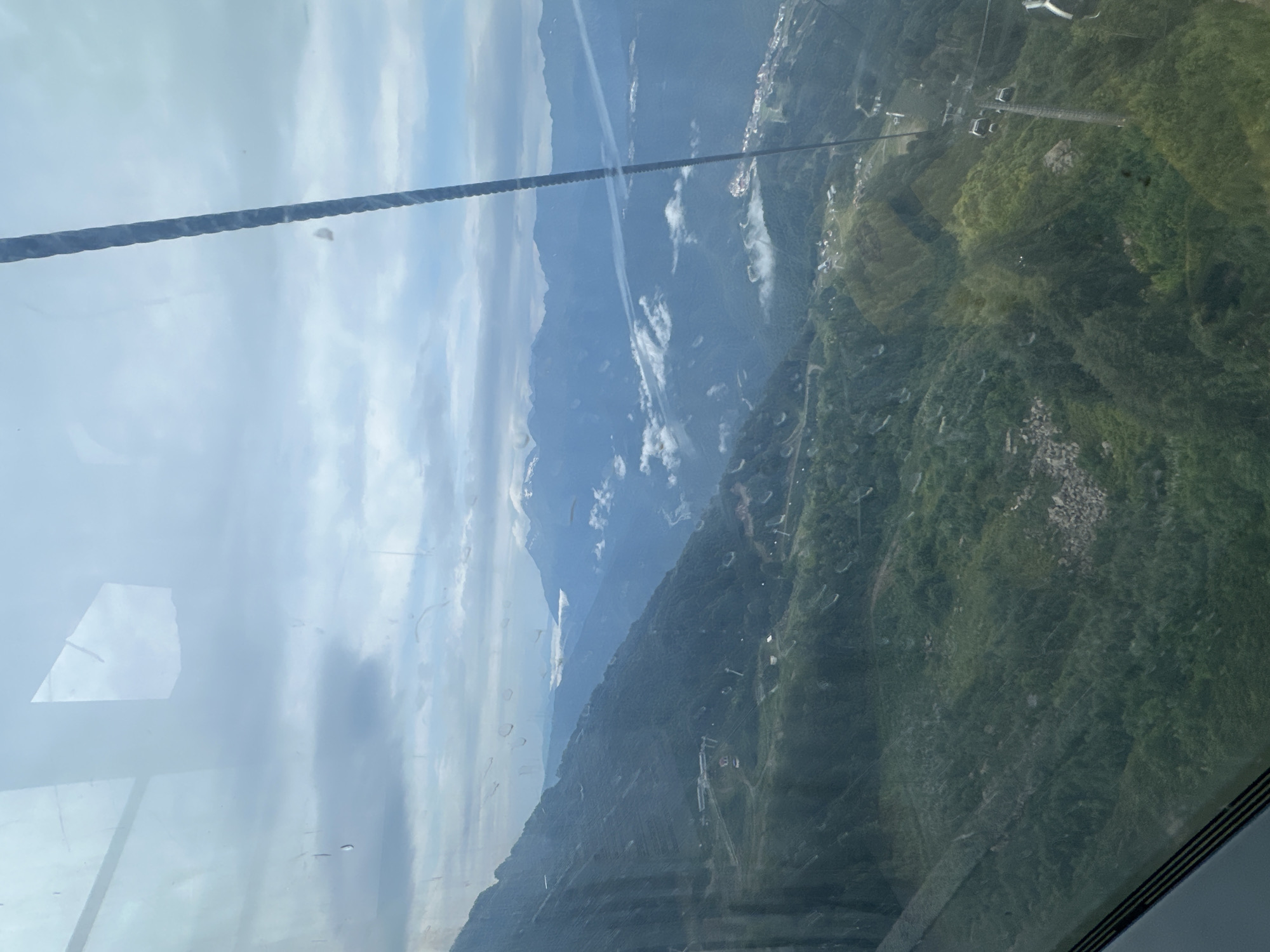 This is a photograph taken from a cable car in Estosadok, Russia, looking out over a vast mountainous landscape. The view is partially obscured by the window of the cable car, which shows reflections and streaks.
The foreground is dominated by the green, forested slopes of mountains, with patches of exposed rock and some evidence of human development, such as buildings and what appear to be ski lift infrastructure with gondolas. A prominent cable stretches diagonally across the frame, part of the cable car system.
In the midground, more mountains recede into the distance, shrouded in a light mist or clouds. These mountains are layered in shades of blue and grey, with some peaks capped with snow or bright white clouds. Rivers or valleys are visible in some of the lower-lying areas between the peaks.
The sky is overcast with a mixture of grey and white clouds, suggesting a cool or temperate day. The overall impression is one of a grand, natural vista with human elements integrated into the environment. The specific location in Estosadok, Russia, is consistent with a mountain resort area that utilizes cable car transportation for access to higher elevations.