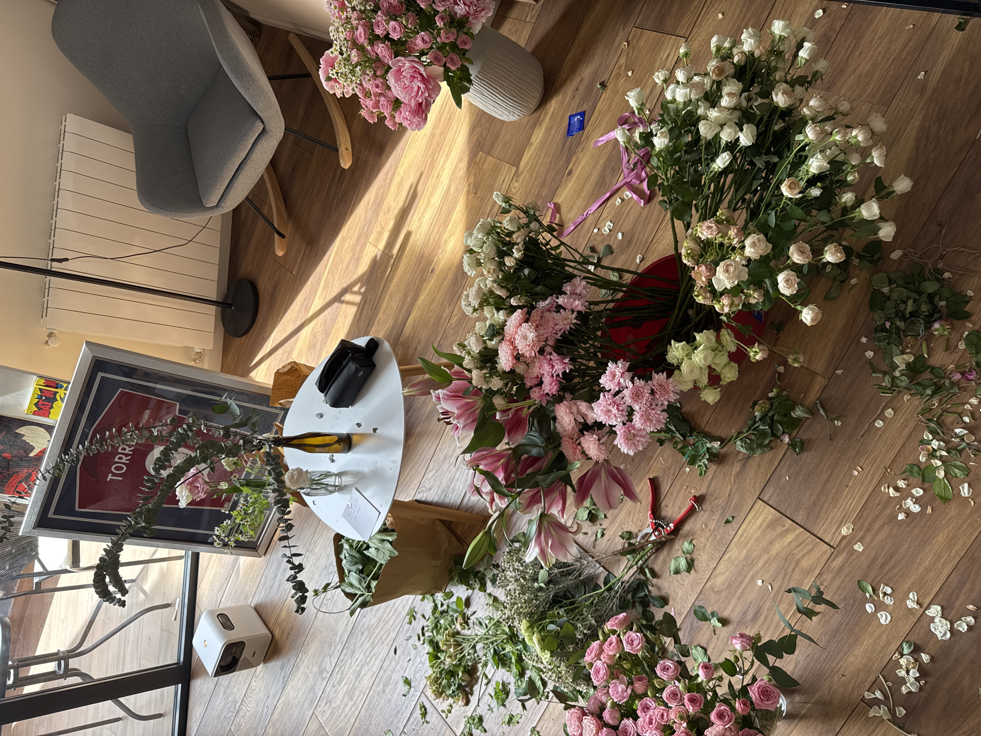 The image displays an indoor scene, likely a living space or studio, filled with a vibrant assortment of flowers being arranged or processed. Bright sunlight streams into the room, casting distinct shadows across the light brown wooden floor, indicating daytime.
The primary subjects are various flowers, predominantly in shades of pink and white. Large bunches of white roses, small pink roses, pink lilies, and pink chrysanthemums are scattered across the floor. Some flowers are in vases: a tall, white ribbed vase holds a lush arrangement of pink peonies and small pink roses, while a clear glass vase on a white round table contains eucalyptus and other greenery. A red bucket also holds a large quantity of white roses and pink chrysanthemums. Scattered petals and trimmed leaves on the floor, along with a pair of red-handled pruning shears, suggest ongoing floristry work.
Other objects in the scene include a grey upholstered rocking chair with wooden and black metal legs, a white round table, and a black floor lamp. A large framed picture, possibly a sports jersey or poster with the word "TORRE" partially visible, leans against a wall. On the table, there is a black case bearing the "DJI" logo and a yellow-brown bottle. A small white projector-like device rests on the floor near the table, and a small blue rectangular object lies on the wooden floor. The overall setting appears clean and organized, despite the creative mess of flower preparation.