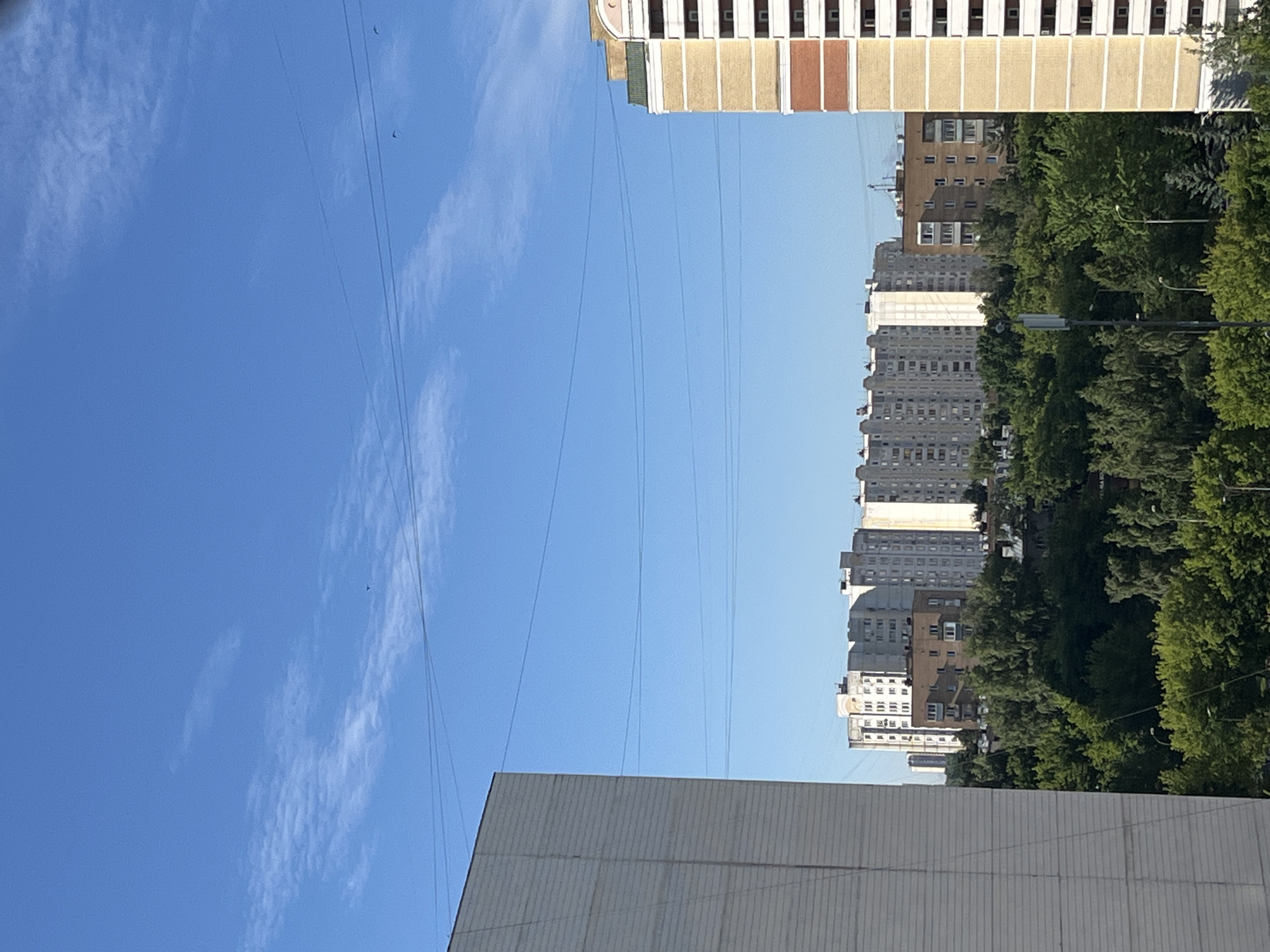 The image captures a scene of a residential area in Moscow, Russia, under a clear blue sky with scattered wispy clouds. The perspective is vertical, with the sky occupying the left half of the frame and buildings and trees on the right. Several tall apartment buildings, characteristic of Soviet-era architecture, are visible. Some are lighter in color with a grid of windows, while others are darker brick or concrete structures. One building at the top is distinct with its cream-colored facade and colorful brick accents, suggesting it might be older or more a modern renovation.
A dense line of lush green trees runs along the base of the apartment buildings, suggesting a park or tree-lined street. Power lines crisscross the sky, creating a network of horizontal lines against the blue. In the foreground, a portion of another building, likely a lower-rise apartment or industrial structure, is visible at the bottom, with a textured grey facade.
The time of day appears to be daytime, likely late morning or afternoon, given the bright sunlight casting shadows and illuminating the scene. The weather is fair and clear. There are no people visible in the frame, and the overall mood is one of urban tranquility. No text is discernible in the image.