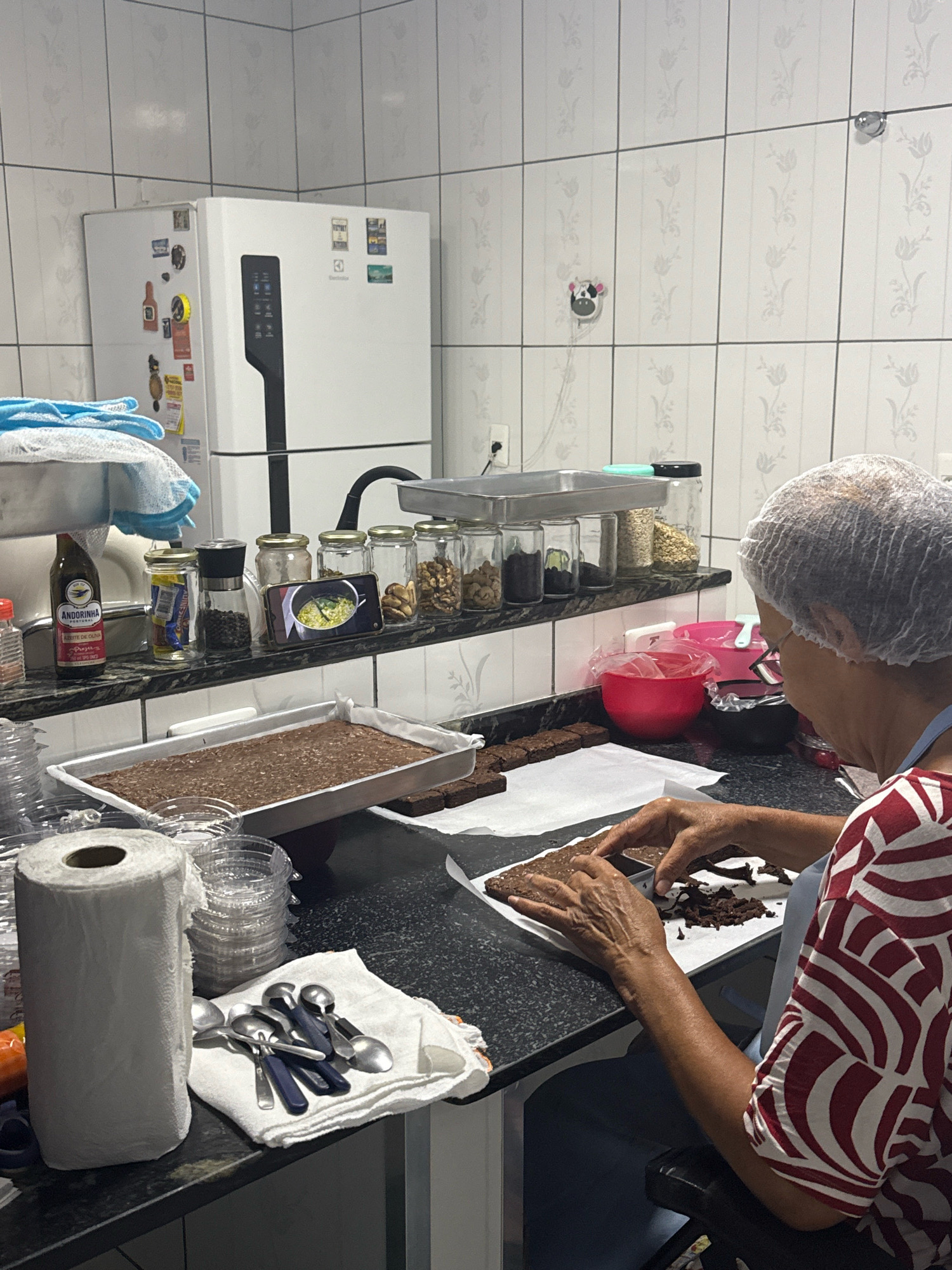 In Nilópolis, Brazil, a person wearing a hairnet and a red and white patterned shirt is seated at a kitchen counter, actively preparing brownies. The person is focused on cutting brownies with their hands, which are resting on a sheet of parchment paper. Scattered chocolate pieces are visible on the paper, indicating the cutting process. In front of the person is a pan containing a large, un-cut brownie. The kitchen environment features a white refrigerator with magnets on its door, a shelf stocked with jars of nuts and spices, and various kitchen utensils, including a roll of paper towels and spoons, neatly arranged on the counter. The background consists of tiled walls.