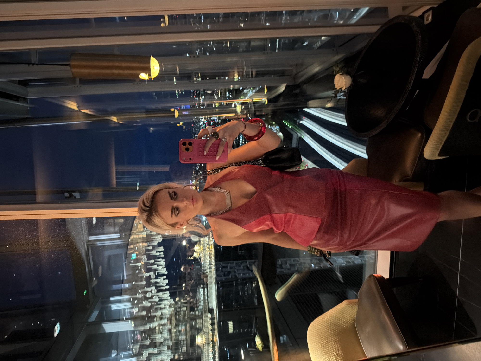 A blonde woman takes a mirror selfie in a restaurant with a view of London at night. She is wearing a red leather halter top and skirt, a statement necklace, hoop earrings, and bracelets. She holds a pink phone in her right hand. In the background, the cityscape is visible through a large window, along with the interior of the restaurant, featuring chairs and tables. The lighting is dim, with some decorative lights in the foreground and the city lights in the distance.