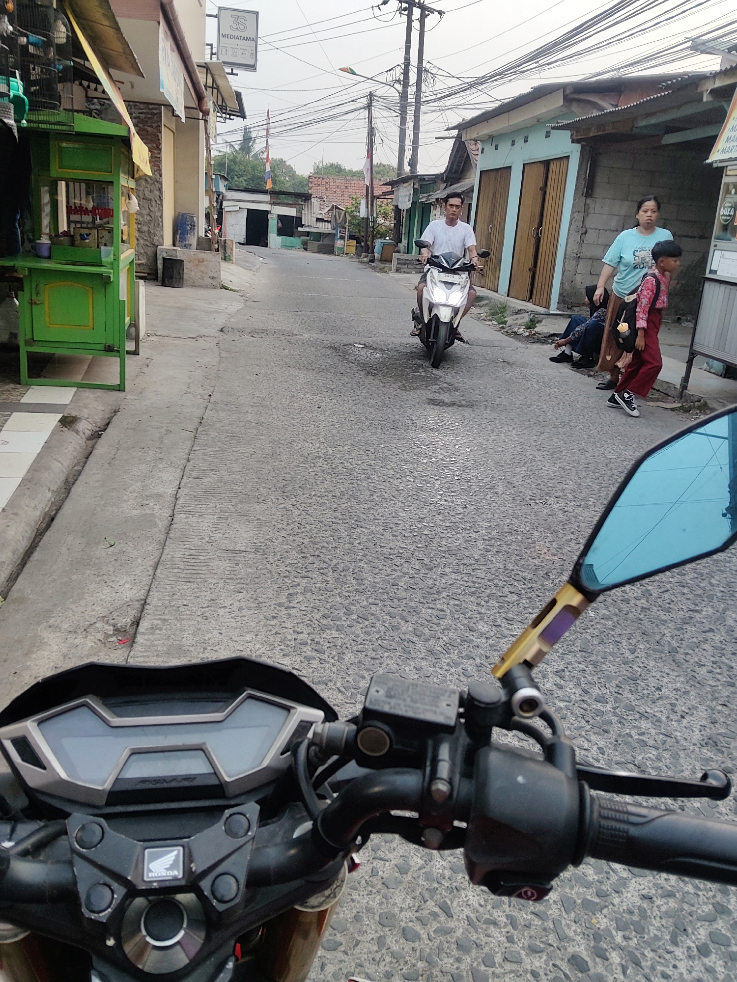 The image depicts a street scene in Mekarmukti, Indonesia, viewed from the perspective of someone riding a motorcycle. In the foreground, the motorcycle's handlebars and dashboard are visible, with a blue side mirror to the right. On the street, a man wearing a white t-shirt is riding a white motorcycle. To the right of the road, a woman in a blue t-shirt and a child in a red and pink outfit are walking. Another person is seated on the ground behind them. Along the left side of the street, a green food cart with yellow trim is present, and further down the road, buildings with corrugated metal roofs are visible. The sky is overcast.