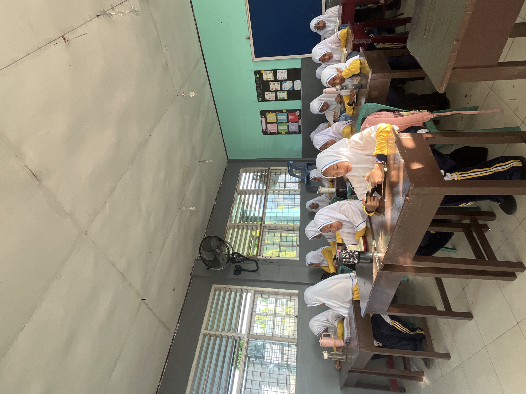 The image shows a classroom with several young female students wearing hijabs and yellow tops, sitting at wooden desks. They appear to be engaged in reading or studying. The classroom has windows with horizontal blinds, a ceiling fan, and posters on the wall. The overall impression is of a typical school environment in Indonesia, possibly Lhokseumawe.