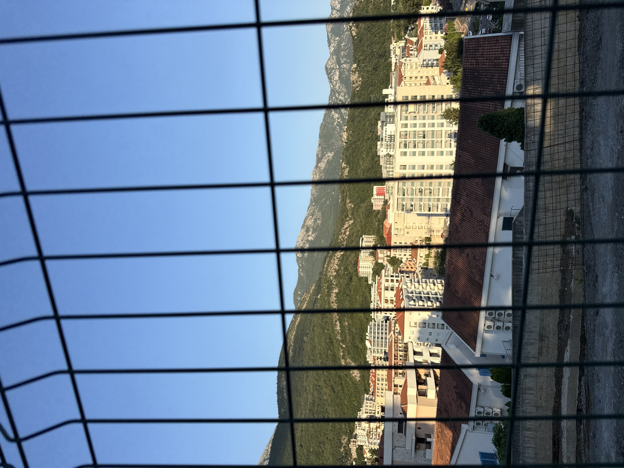 The image shows a view of the city of Rafailovići, Montenegro, from behind a wire fence. In the foreground, the dark green wire fence obstructs the view. Beyond the fence, a cityscape with several buildings of varying heights and architectural styles is visible. Many of these buildings have white or light-colored facades and red-tiled roofs. Some buildings appear to be apartment complexes or hotels, characterized by multiple floors and balconies. In the background, lush green mountains rise steeply, covered in dense vegetation. The sky is a clear, bright blue, indicating a sunny day. The lighting suggests it might be late afternoon or early morning due to the soft shadows. There are no people or vehicles clearly visible in the frame.