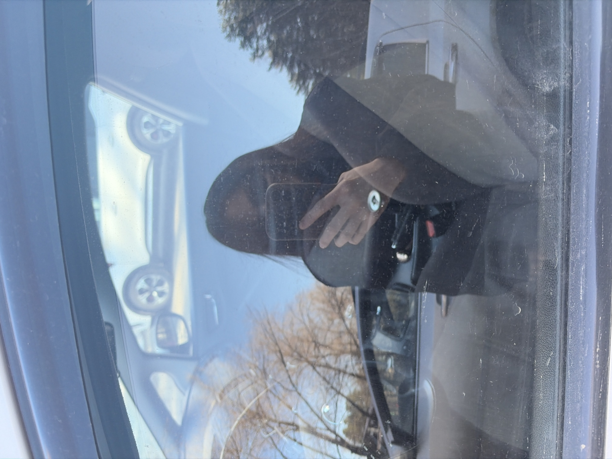 This media file captures a complex reflection within a car window, likely a windshield or side panel, in what appears to be Chaikovsky, Russia. The image is presented rotated 90 degrees clockwise from a typical upright perspective.
The primary subject is a partial reflection of a person, presumably the photographer, holding a dark-colored smartphone. Their left hand, wrist, and forearm are visible, featuring a round-faced watch with a light-colored strap. The silhouette of their head and shoulder is also discernible.
The reflections blend elements from both inside and outside the vehicle. Faint reflections of the car's interior, including a dark, textured panel, are visible. The exterior environment reflected shows a bright, clear sky, indicating a sunny day. Bare trees, suggestive of late autumn or winter, along with some evergreen foliage, are clearly reflected, aligning with the provided location context of Chaikovsky, Russia. Further external reflections include a five-spoke alloy wheel and potentially a side mirror of another vehicle.
The car window itself appears somewhat dusty or smudged, with visible particles and marks contributing to the diffused quality of the reflections. No discernible text is visible within the image.