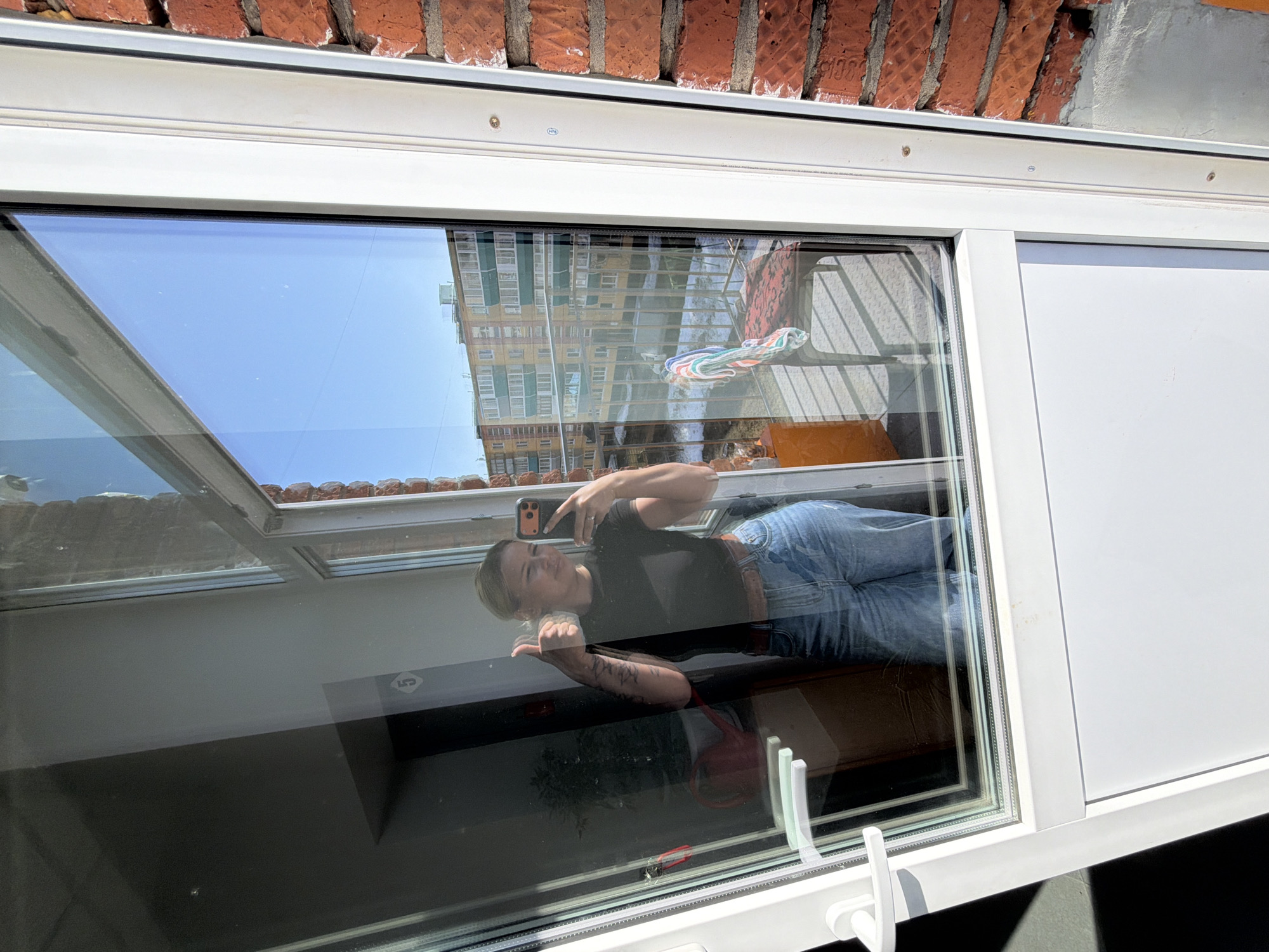 The photograph depicts a woman taking a selfie in front of a window. The window is reflecting the exterior of a building and the sky. The building's facade is visible in the reflection, indicating an urban environment. The woman is wearing a dark top and jeans. The window frame is white, and the building exterior appears to be made of red brick. Without additional information, pinpointing the exact location in Sarapul, Russia, is impossible.