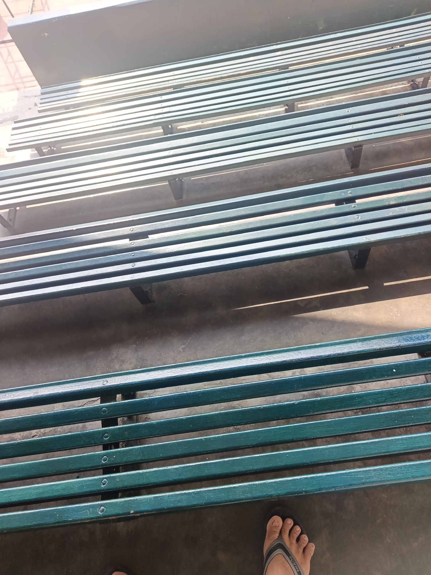 The image displays multiple rows of dark green, slatted benches, possibly in a public seating area. The benches appear to be made of metal or wood and are arranged in tiers. Sunlight casts distinct shadows on the concrete ground beneath the benches, suggesting an outdoor setting. In the lower right corner, a person's left foot, wearing a dark grey flip-flop with a lighter grey and black strap, is visible, indicating a person's presence in the scene. The overall environment appears to be a public space in East Jakarta, Indonesia, given the context provided.