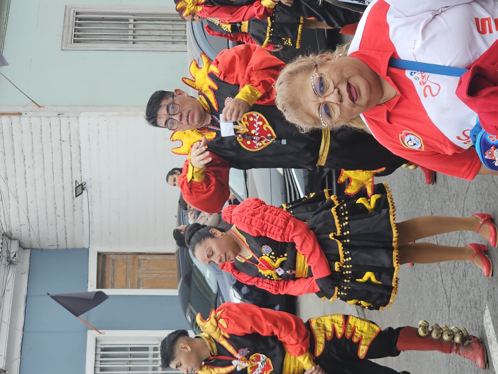 The image depicts a street scene in Iquique, Chile, featuring individuals in traditional festive attire. In the foreground, a woman wearing glasses and a red and white shirt with a blue strap across her shoulder is looking towards the camera. Behind her, a group of people are dressed in elaborate costumes that include black and red outfits adorned with yellow accents. One man in this group is wearing a black garment with red sleeves and yellow flame-like embellishments on the shoulders and chest, holding a white object in his hand. To his left, a woman in a similar costume, featuring a black skirt with red ruffled sleeves, stands with her eyes closed. Another man on the far left is also dressed in a matching costume. The background shows a street with parked cars and buildings, one of which has a blue facade and a wooden door. A black flag is visible mounted on the building. The overall atmosphere suggests a cultural event or parade.