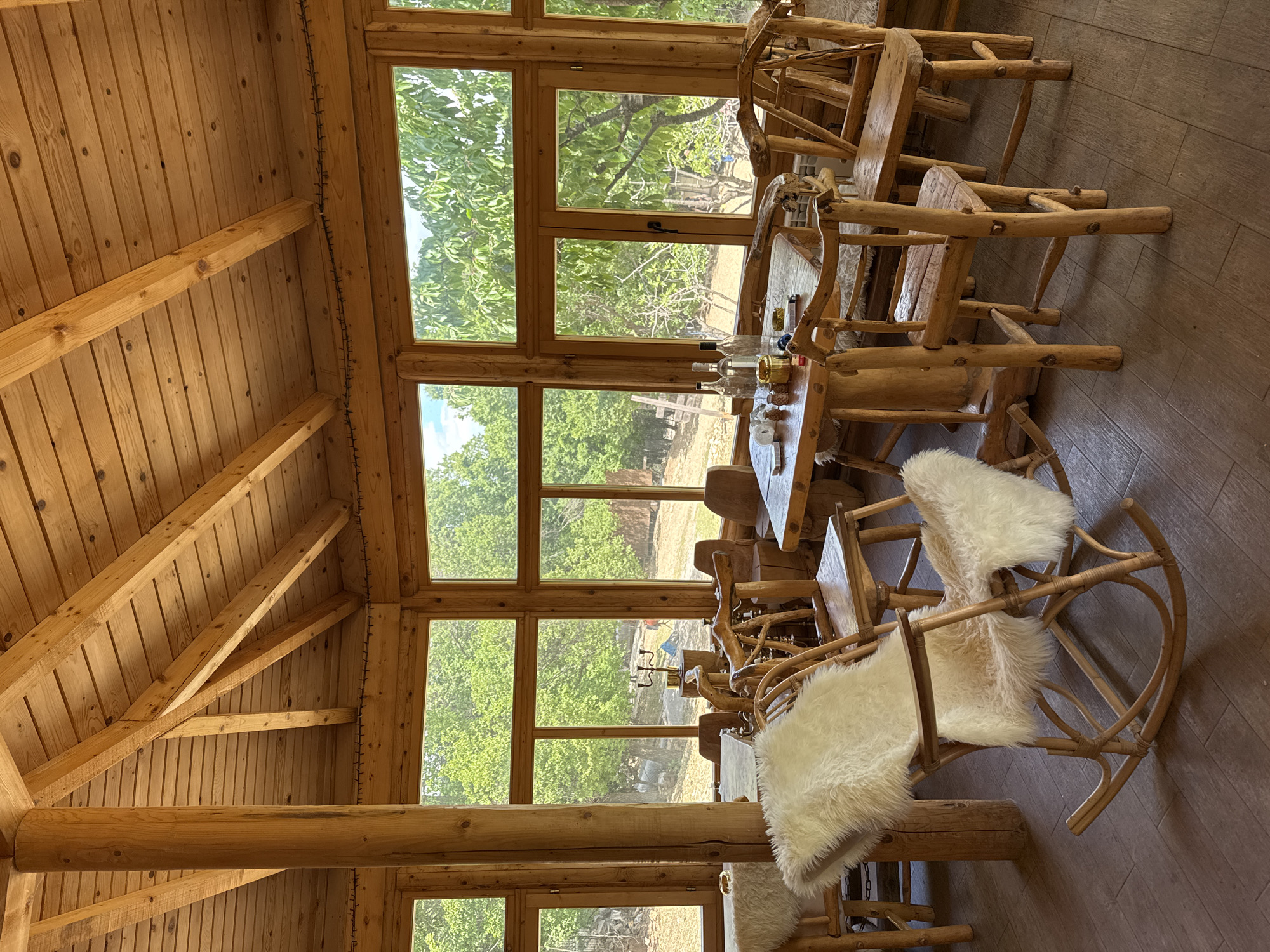 The image depicts the interior of a rustic wooden cabin, likely located in a rural area around Gornje Mrke, Montenegro. The cabin's walls and ceiling are constructed of light-colored wood, creating a warm, natural ambiance. Large windows provide an expansive view of the outside, revealing lush green trees and a glimpse of the landscape.
A wooden table is centrally positioned, surrounded by unique, handcrafted wooden chairs, some adorned with white fur seat covers. The table appears to be set, with bottles, glasses, and other items suggesting a recent or upcoming meal. The overall impression is one of a comfortable, secluded retreat, blending natural materials with an appreciation for the surrounding environment.