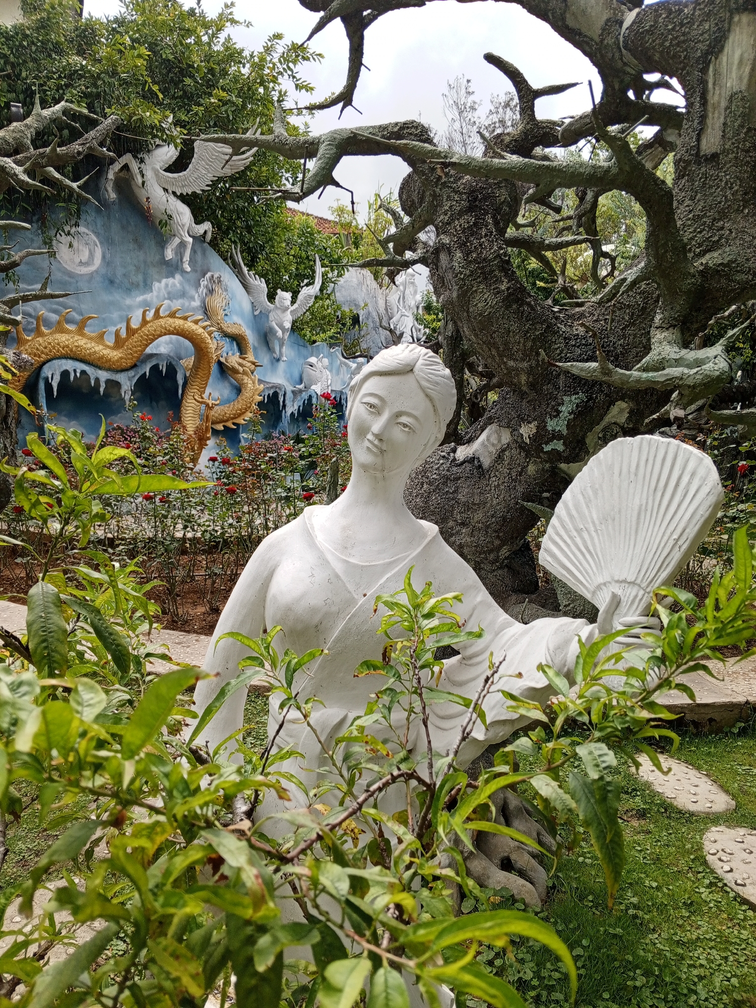 The image shows a white stone statue of a woman holding a fan in a garden setting. Behind the statue, a mural depicts a golden dragon and white winged creatures, possibly mythical beings, against a blue, icy backdrop. The garden features lush green foliage in the foreground, with a large, gnarled tree trunk to the right. Red roses are planted in beds behind the statue. The overall scene suggests an artistic or cultural attraction, potentially a garden or temple in Da Lat, Vietnam, known for its many gardens and floral displays.