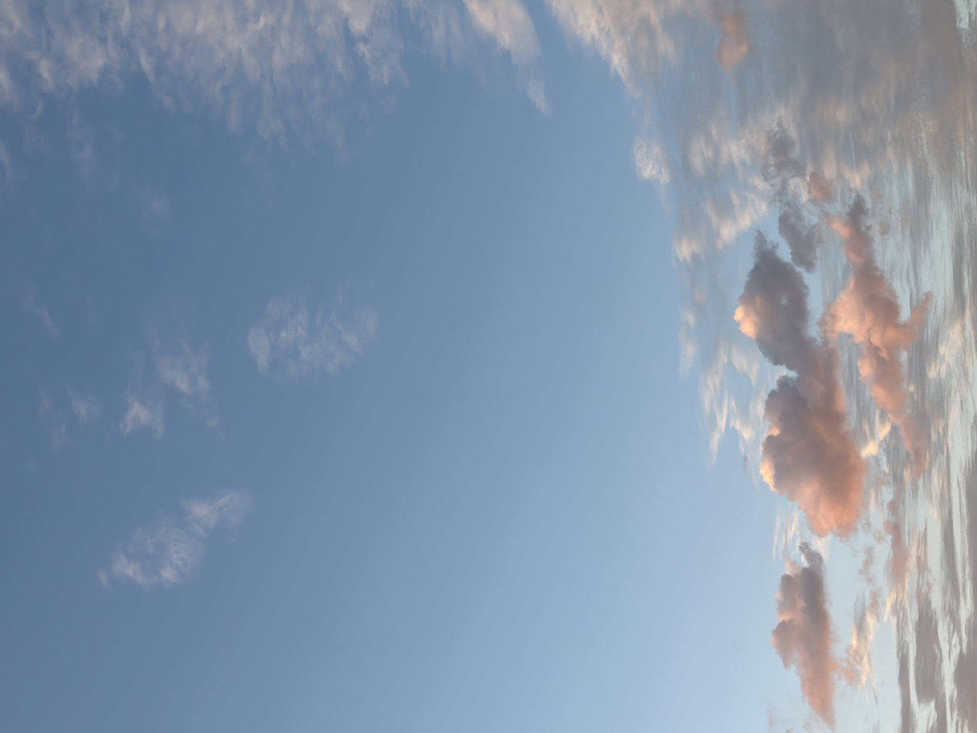 This is a vertical, eye-level, medium shot of a sky filled with clouds at sunset. The sky is a pale blue, fading to a lighter shade near the horizon. Wispy white clouds are scattered across the upper portion of the image, while larger, more voluminous clouds with touches of pink and orange are visible in the lower half. The lighting suggests a soft, diffused light, typical of either sunrise or sunset. The scene is devoid of any human presence, structures, or terrestrial elements, focusing solely on the atmospheric conditions. The image does not depict any specific identifiable landmarks or context that would place it in Узюково, Russia, or any other specific geographic location.