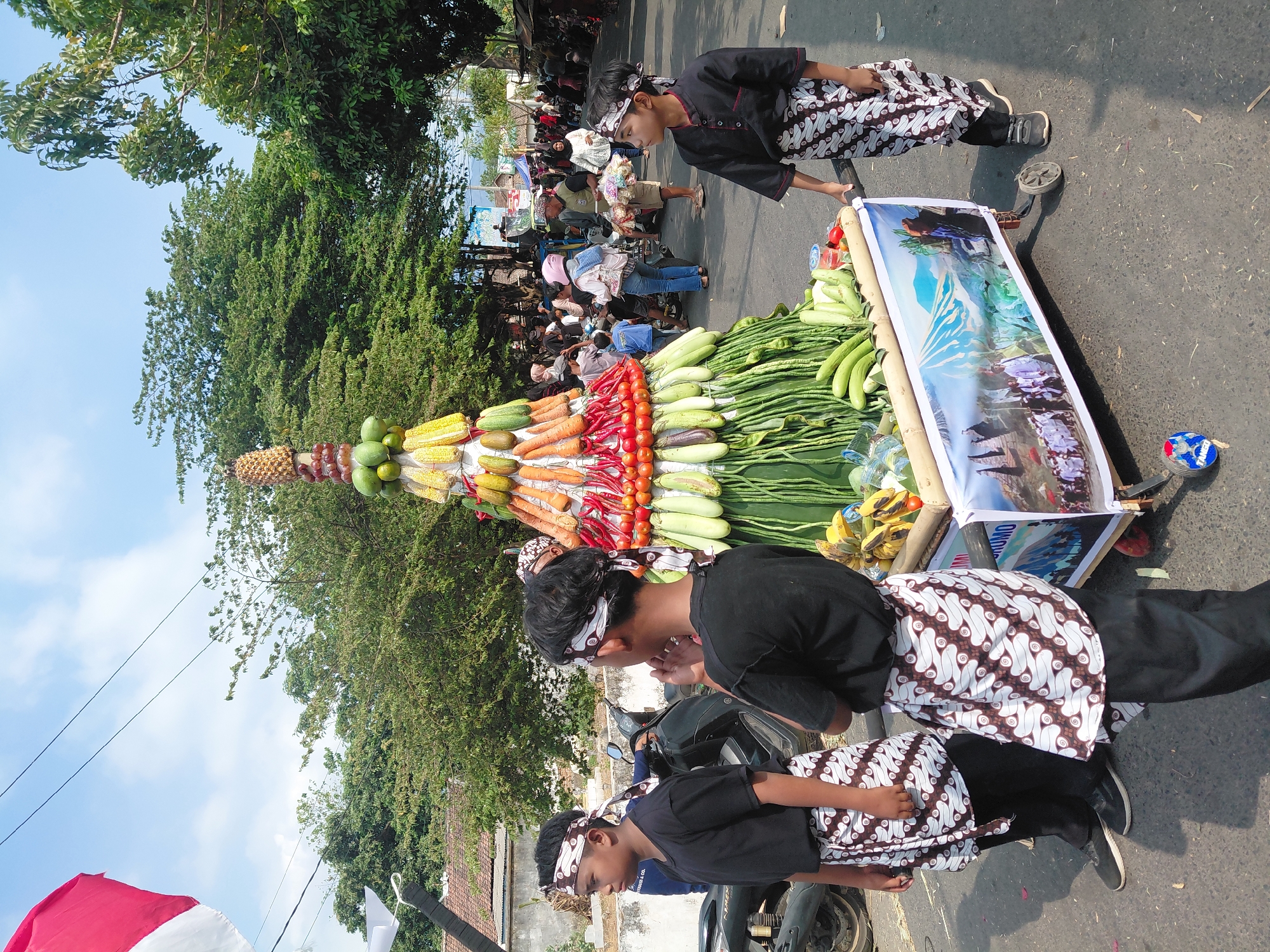 In the city of Bima, Indonesia, a vibrant street scene unfolds, featuring a procession centered around a towering, conical arrangement of fruits and vegetables. The structure, adorned with corn, carrots, chilies, tomatoes, and various green vegetables, sits atop a wheeled cart, displaying a banner with images of a mountain and people in procession. Two young boys, dressed in black shirts, black pants, and patterned sarongs, stand beside the cart, appearing to be participants or observers. Their heads are adorned with matching patterned headbands. In the background, a crowd of people, some on motorcycles, are gathered, suggesting a festive or cultural event. The environment is a paved street lined with trees and houses, under a bright blue sky with scattered clouds. An Indonesian flag flutters in the upper left corner of the image.