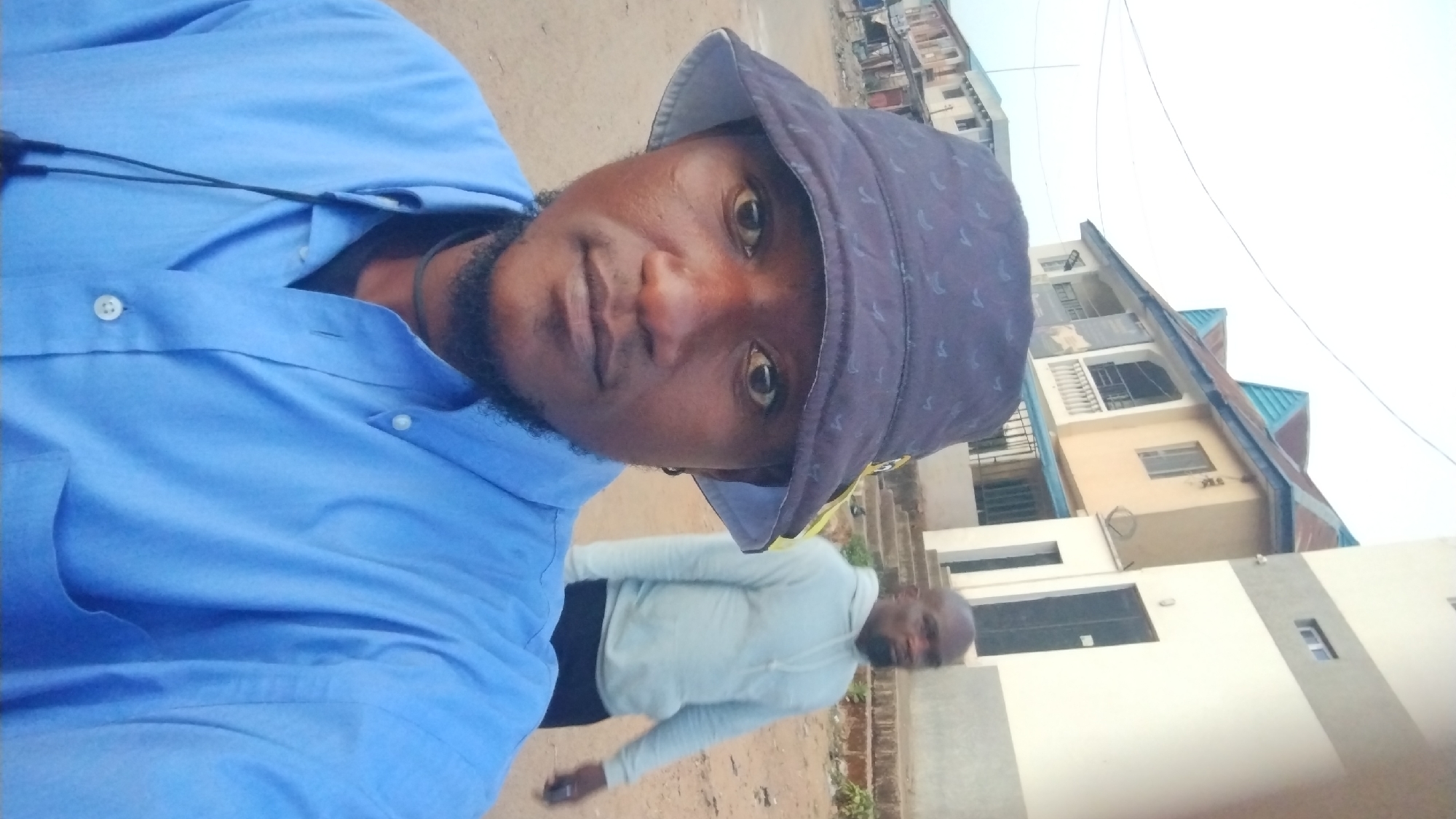 A photograph, likely a selfie, depicts a man wearing a blue button-down shirt and a dark-colored bucket hat. He has dark skin, a beard, and appears to be outdoors during daylight. Behind him, another man in a light-colored long-sleeved shirt and dark pants is partially visible.
The background includes buildings, suggesting an urban or suburban setting. The architecture of the buildings appears to be in the style commonly found in the city of Jos, Nigeria, given the color of the walls and roofs, as well as the building materials, although the specific location cannot be verified with certainty based solely on this image. The ground appears to be dirt or a similar unpaved surface. Overhead electrical lines are visible.