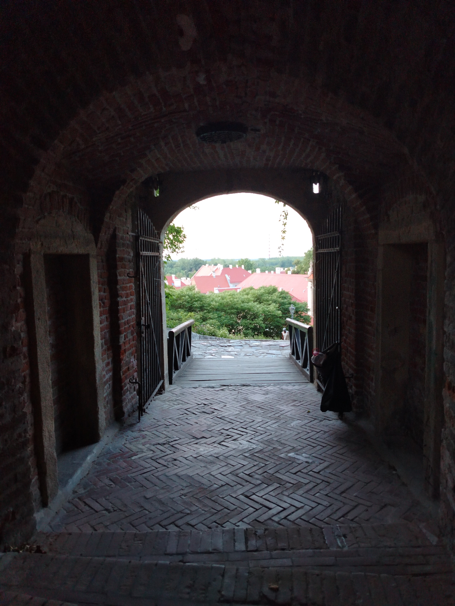 The image captures a perspective looking out from a brick-lined tunnel, likely within the Petrovaradin Fortress in Petrovaradin, Serbia. The tunnel has a vaulted brick ceiling and two arched doorways on either side of the entrance. A paved walkway made of herringbone-patterned bricks leads through the tunnel and out onto a wooden bridge. The bridge has a dark wooden railing with a crisscross design. Beyond the bridge, a view of a cityscape with red-tiled roofs and lush green trees is visible. The sky is overcast, suggesting it is either early morning or late evening. A dark-colored object, possibly a bag or coat, is hanging on the right-side gate of the tunnel.