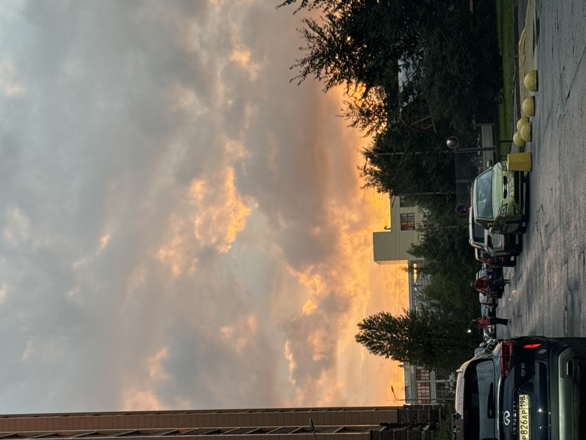 The image depicts a street scene in Sinyavino, Russia, during what appears to be sunset. A tall, modern apartment building is visible on the left side of the frame, its windows reflecting the sky. The sky is filled with dramatic, fiery orange and pink clouds, indicative of a colorful sunset. In the foreground, several cars are parked along the wet asphalt road, suggesting recent rainfall. A dark-colored Infiniti SUV with a license plate reading "P826AP 198 RUS" is prominently featured on the left. Further down the street, a person is walking, and another individual is riding a bicycle. Green trees line the sides of the road, interspersed with buildings in the background. Yellow, dome-shaped bollards are placed along the curb. The overall atmosphere is one of an urban environment transitioning into evening under a striking sky.