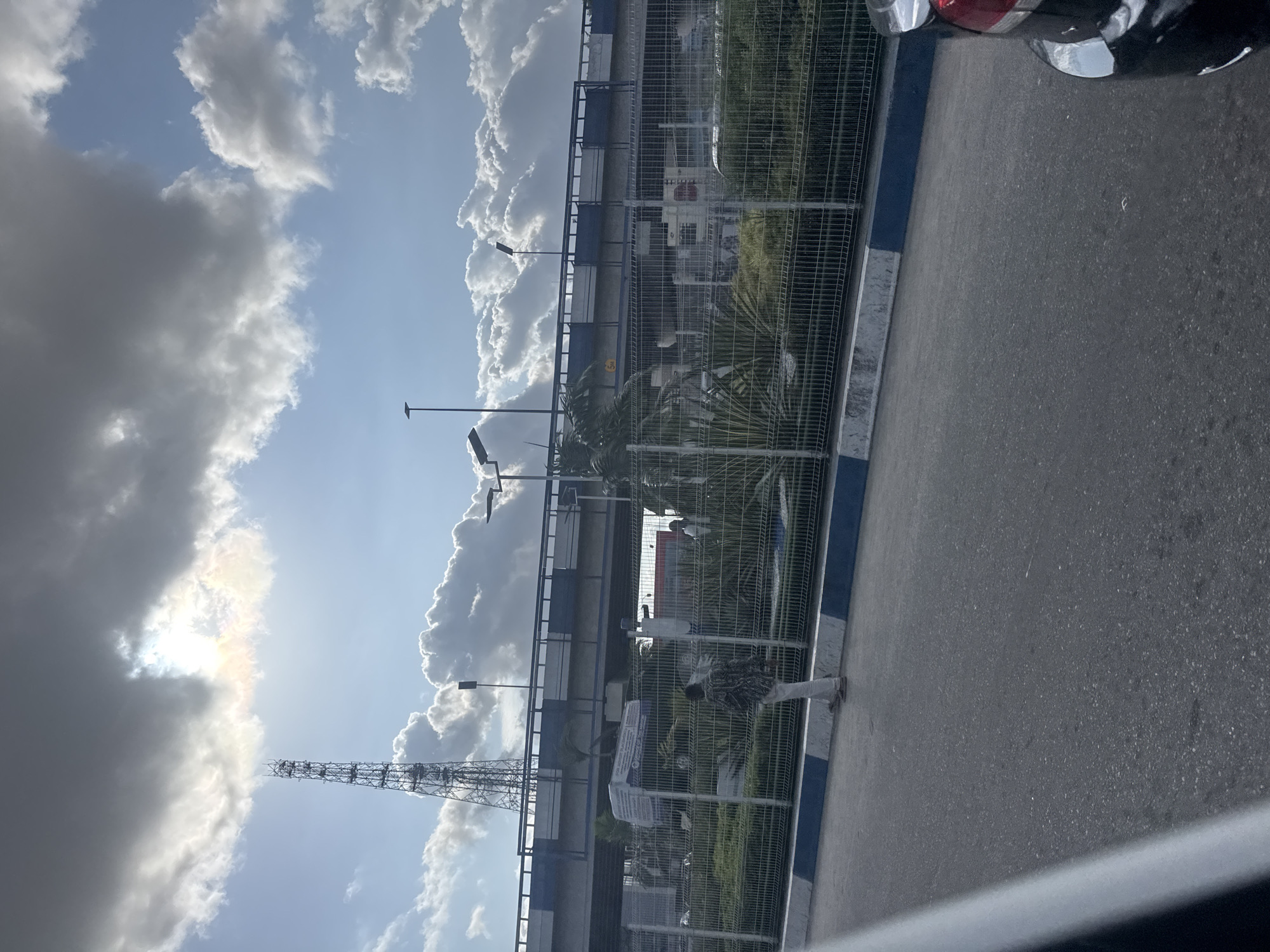 The image depicts a partly cloudy sky over a location likely in Port Harcourt, Nigeria, based on the presence of palm trees and architectural styles common in the region. In the foreground, there is a gray paved area, possibly a road or parking lot, with a blue and white painted curb. A metal chain-link fence separates the paved area from a green area with vegetation. Behind the fence, a building with a red and white exterior is visible. Streetlights extend into the scene above the fence. A lattice tower stands in the distance against the sky. A portion of a car is visible at the top right edge. A person in casual attire can be seen walking within the fenced area.