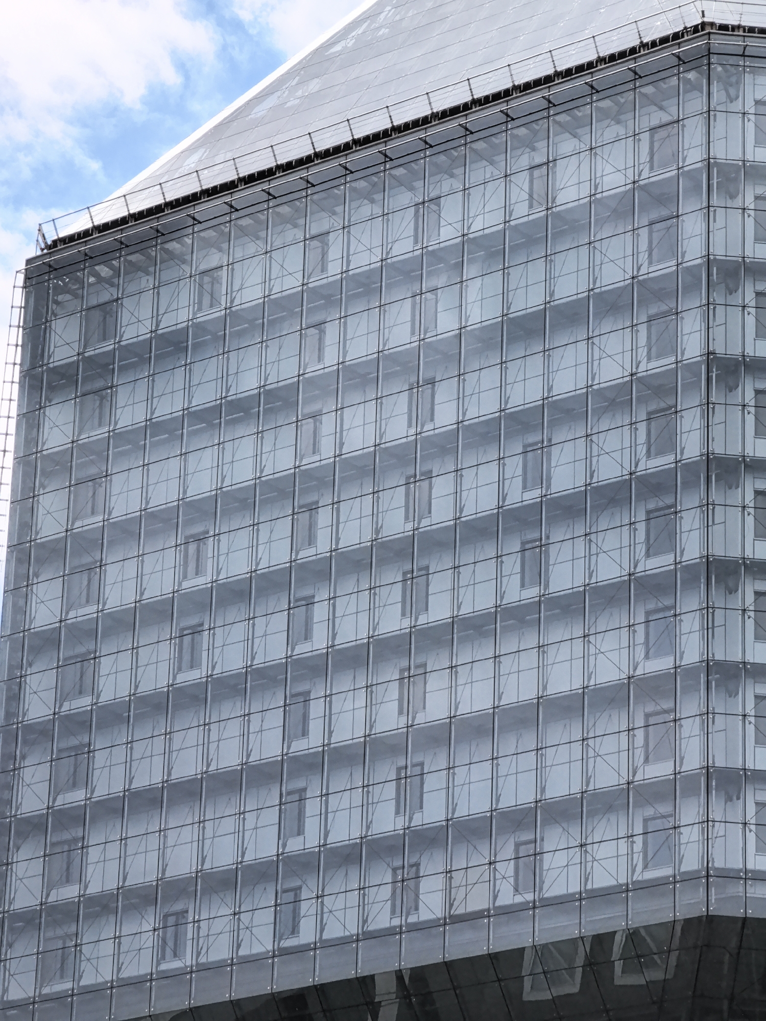 This is a medium shot, eye-level, outdoor photograph of a modern glass-faced building. The building has a distinctive geometric shape, with a large, angled section at the top that appears to be covered in a lighter-colored material, possibly scaffolding or a protective tarp. The main body of the building is covered in a grid of glass panels, framed by a metal structure.
Through the glass, the interior of the building is visible, revealing a uniform pattern of windows and what appear to be balconies or external walkways. The windows are uniformly spaced in a grid, with many of them appearing to be closed. The lighting suggests it is daytime, with some clouds visible in the sky above the building. The overall tone of the image is cool and somewhat monochromatic, due to the glass facade and the overcast sky. There is no visible text in the image. The scene is devoid of people and focuses solely on the architecture. The building appears to be under construction or renovation, given the material covering the upper section and the visible internal structure.