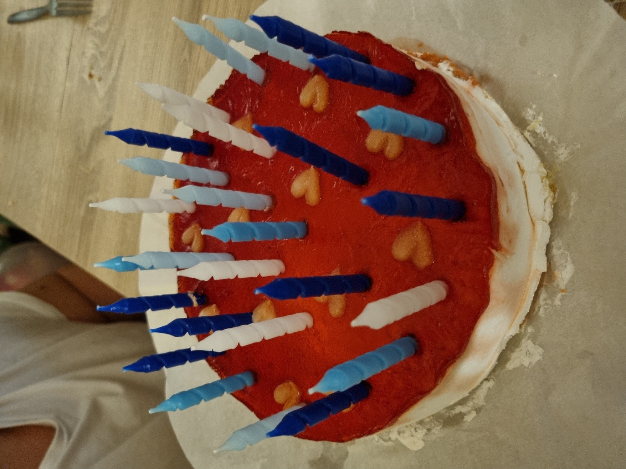 A top-down, slightly angled shot shows a birthday cake with numerous candles, placed on a white parchment paper on a wooden-patterned surface. The cake has a red surface adorned with small, heart-shaped decorations. Candles in blue, light blue, and white are inserted into the cake. In the background, part of a person wearing a white shirt is visible on the left side, along with a metallic fork. The location is not discernible as Saarbrücken, Germany, without additional information.